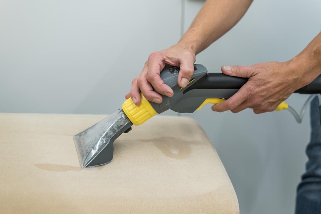Une personne utilise un appareil manuel de nettoyage de tissus d'ameublement avec une poignée jaune et grise pour nettoyer une surface de tissu beige clair.