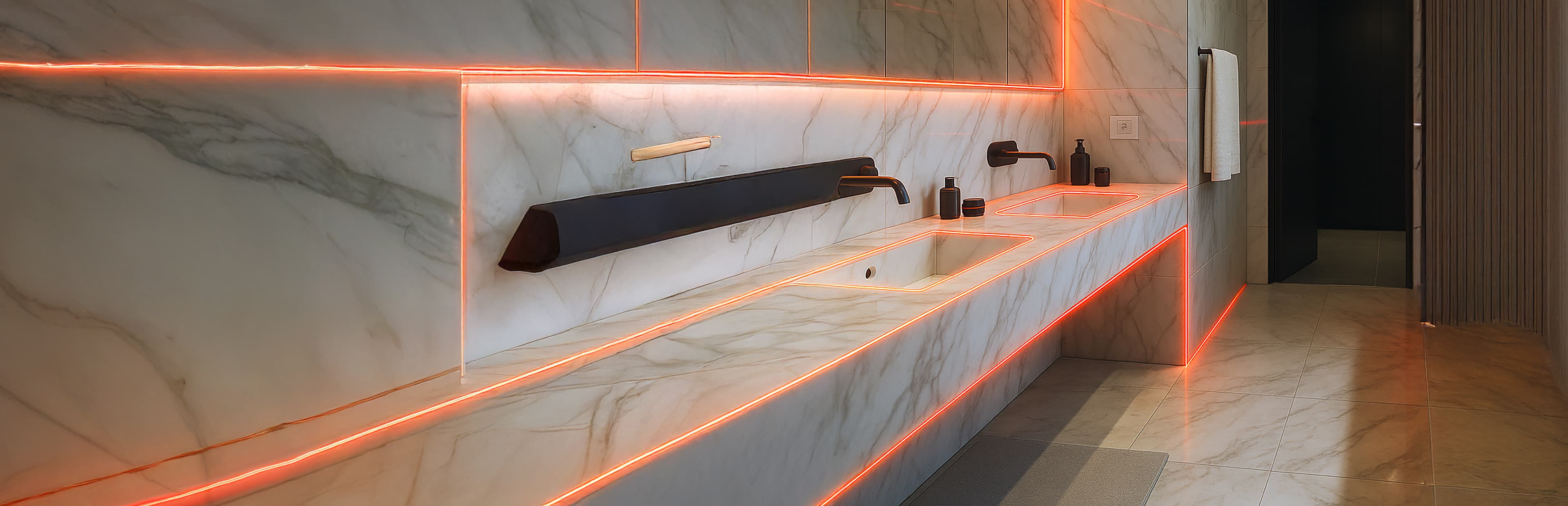 Moderne badkamer met marmeren oppervlakken, dubbele wastafels, zwarte armaturen, zeepdispensers en oranje LED-accentverlichting die de werkbladen en muren accentueert en een strakke, eigentijdse look creëert.