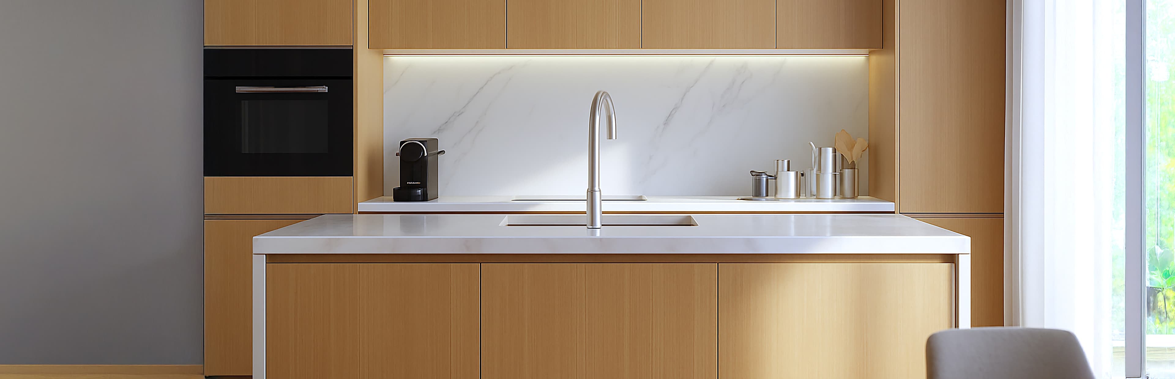 Eine moderne Küche mit hellen Holzschränken, einer weißen Marmorabdeckung und Arbeitsplatte, einem eingebauten Ofen, einer Kaffeemaschine und einer Spüle mit einem hohen Wasserhahn auf einer zentralen Insel. Das Sonnenlicht fällt durch ein Fenster auf der rechten Seite ein.