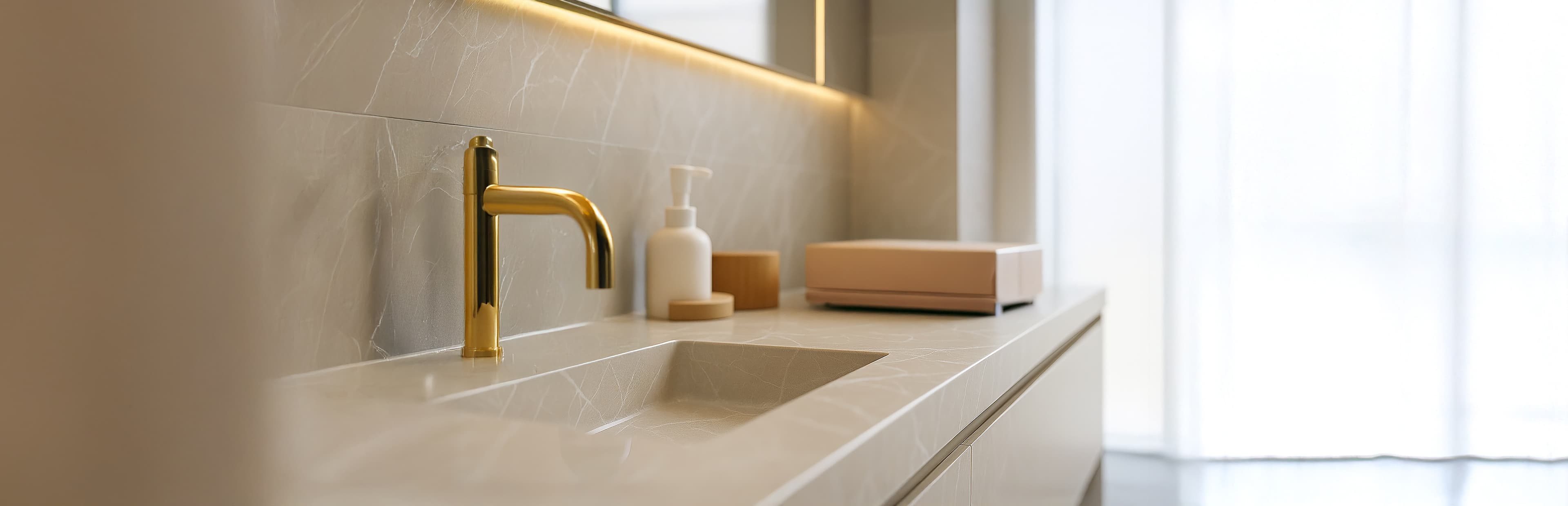 Moderner Badezimmertisch mit einem goldenen Wasserhahn, einem rechteckigen Waschbecken, einem Seifenspender, kleinen Behältern und einer rosafarbenen Schachtel, alles auf einer hellen Marmorfläche. Weiches natürliches Licht fällt durch die transparenten Vorhänge im Hintergrund.