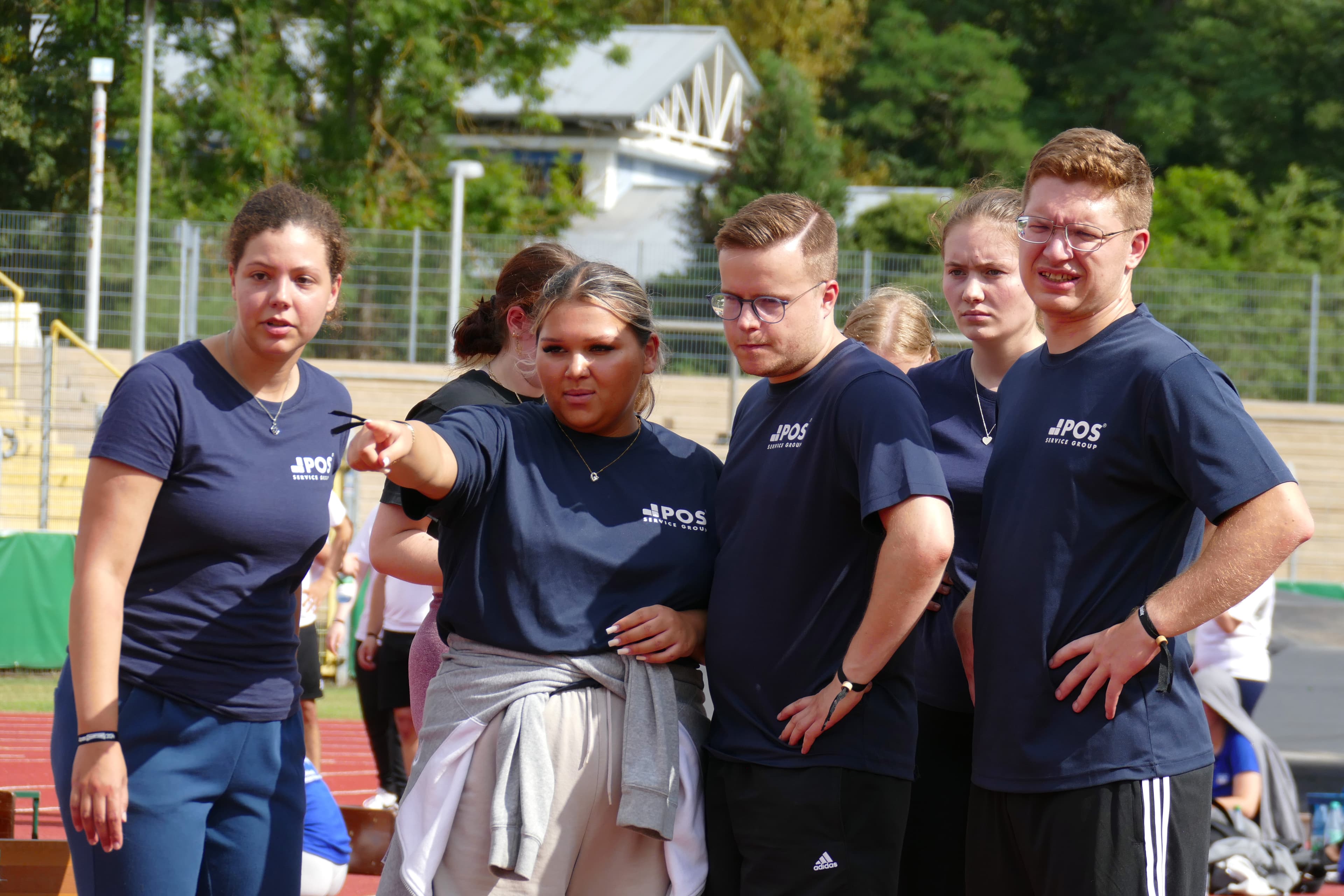 Five young adults in matching navy shirts stand together outdoors on a sports track. One woman points toward something off-frame while the others look in the same direction with focused or concerned expressions.