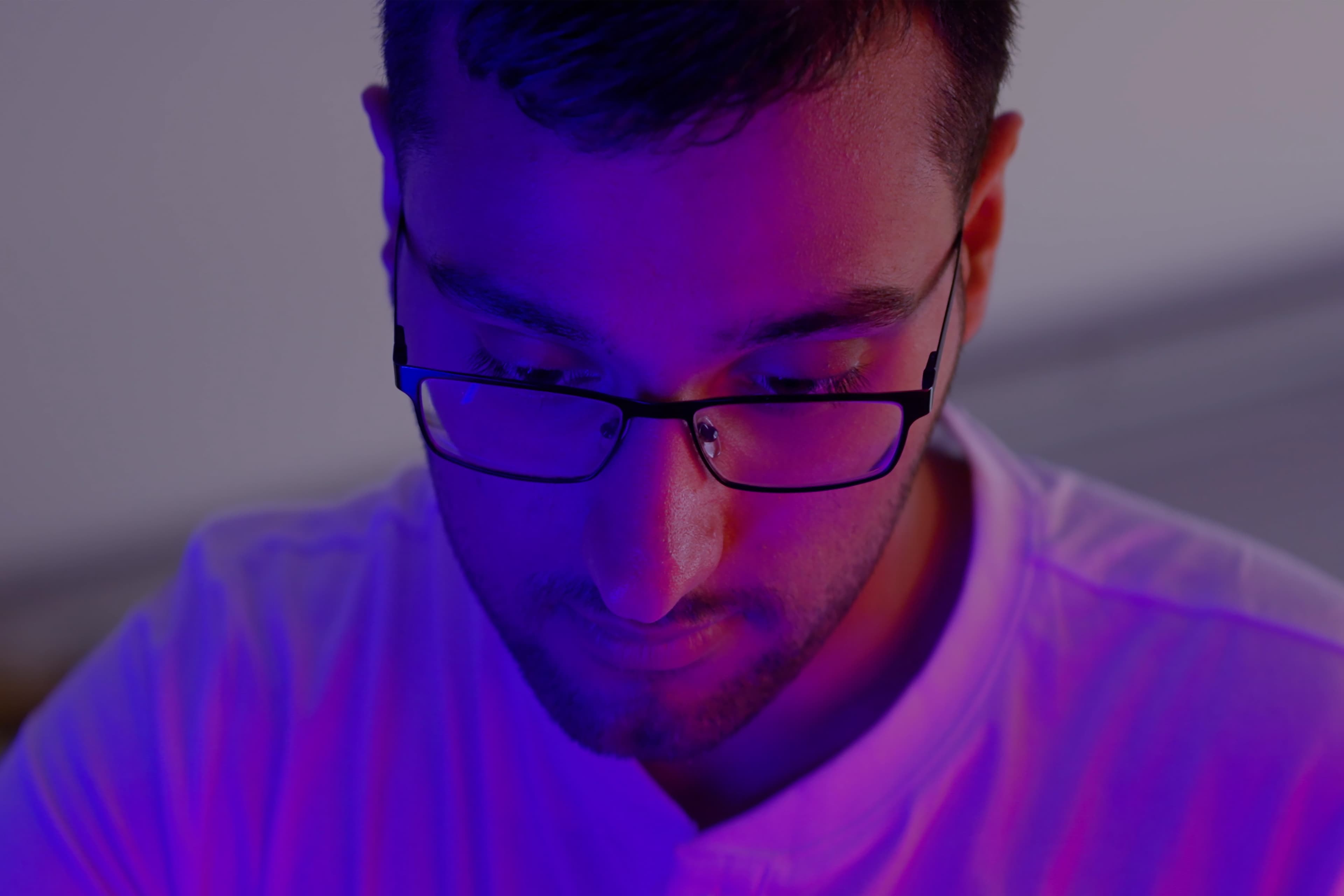 A person wearing glasses and a white shirt looks down, illuminated by purple and blue lighting in a dimly lit room.