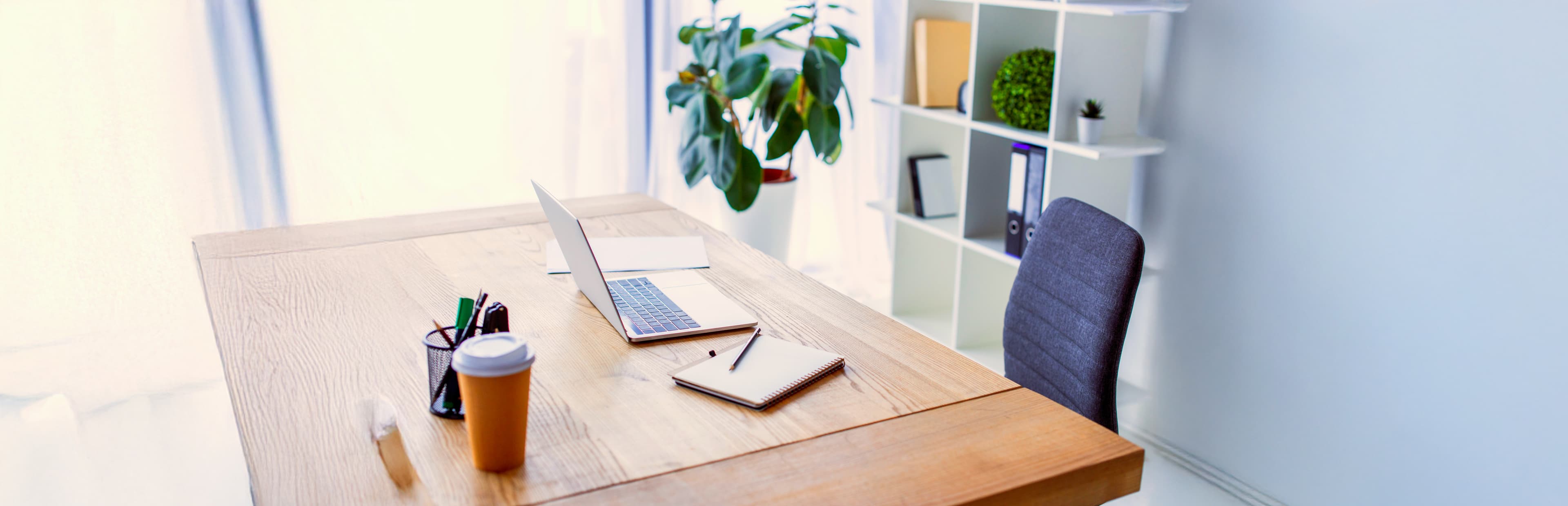 Ein moderner, aufgeräumter Bürotisch mit Laptop, Notizbüchern, Stifthalter und einem Kaffeebecher zum Mitnehmen. Im Hintergrund stehen eine Pflanze und ein weißes Bücherregal mit Akten und Dekorationsartikeln. Helles natürliches Licht erfüllt den Raum.