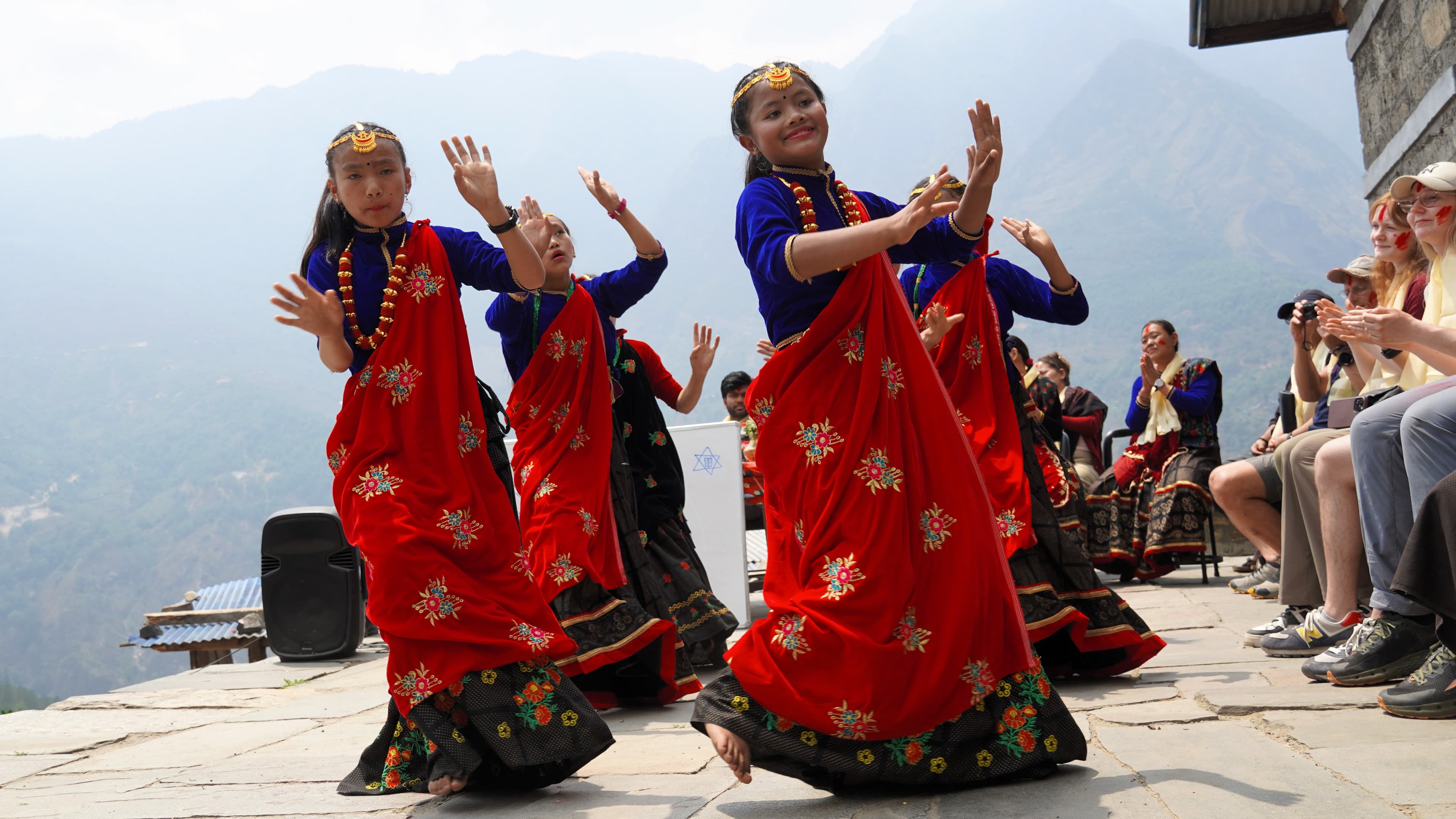 A group of women in vibrant red and blue traditional dresses perform a cultural dance outdoors, with mountains in the background and people watching from the side.