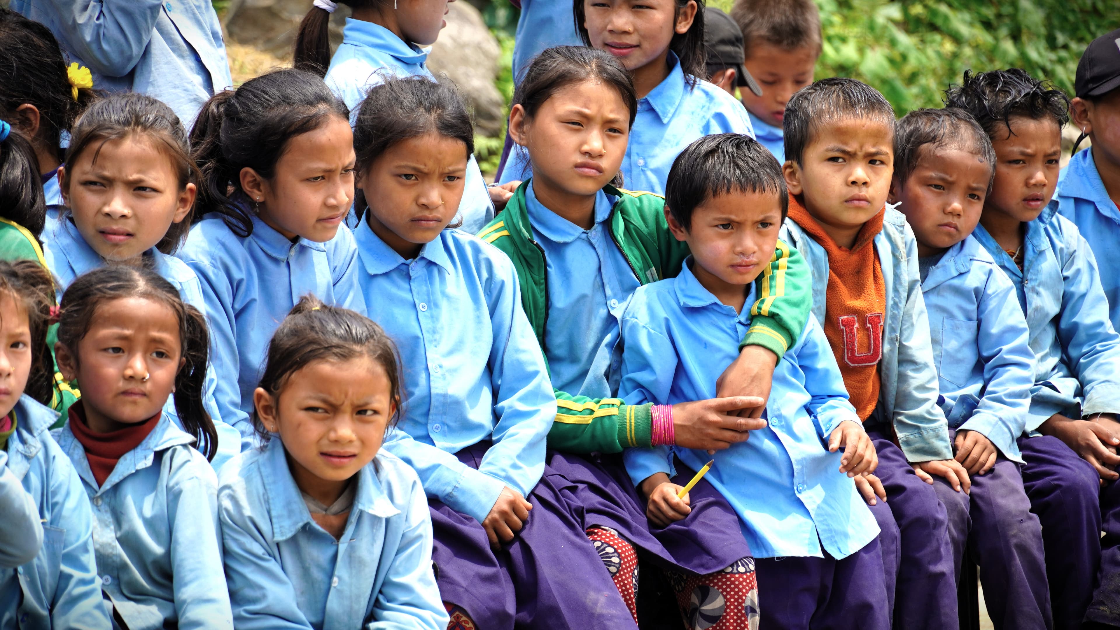 A group of school children sit closely together outdoors, wearing blue uniforms. Most of the children are looking forward with serious expressions, and one girl has her arm around a younger child.