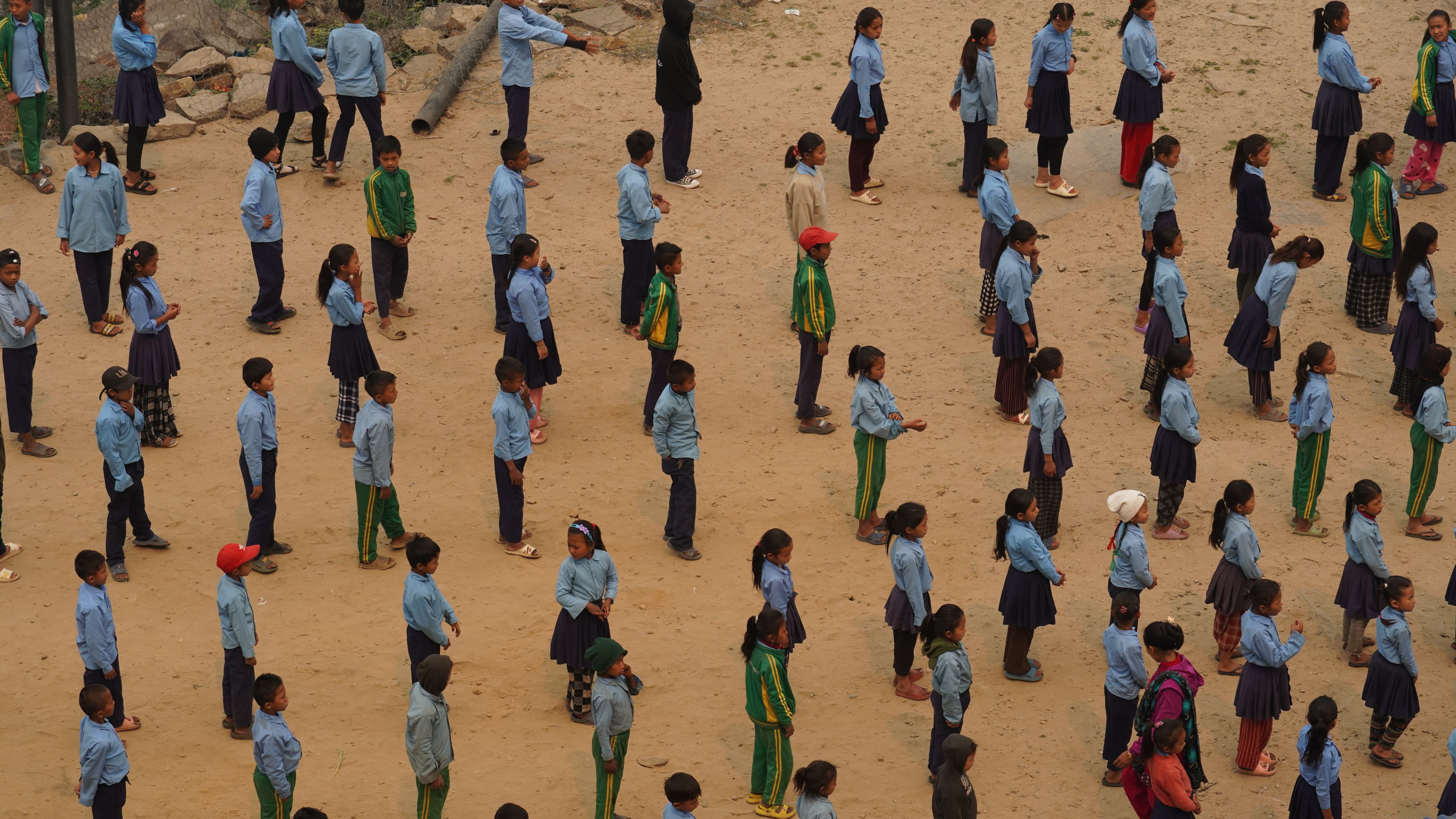 A large group of school children, wearing uniforms of blue shirts and green tracksuits, stand in neat rows on a sandy ground, likely participating in a school assembly or outdoor activity.