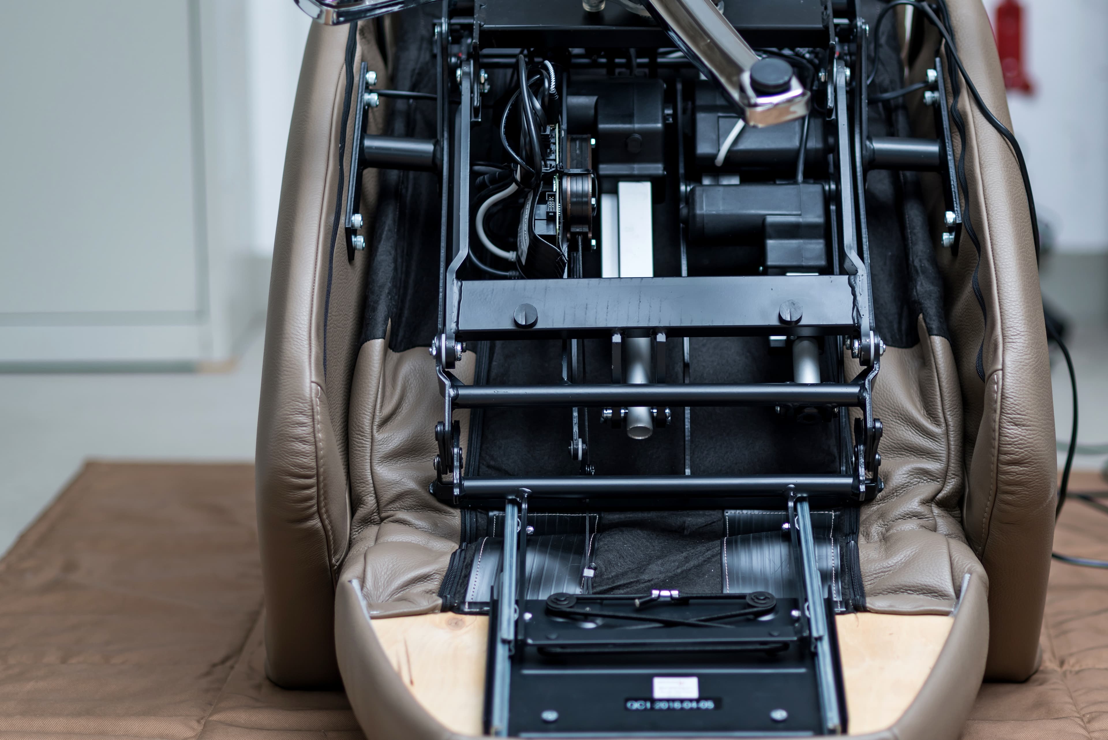 L'image montre le dessous d'un fauteuil inclinable en cuir marron, exposant ses composants mécaniques et électriques, y compris les cadres métalliques, les fils et les pièces mobiles. Le rembourrage est partiellement ouvert pour révéler l'intérieur.