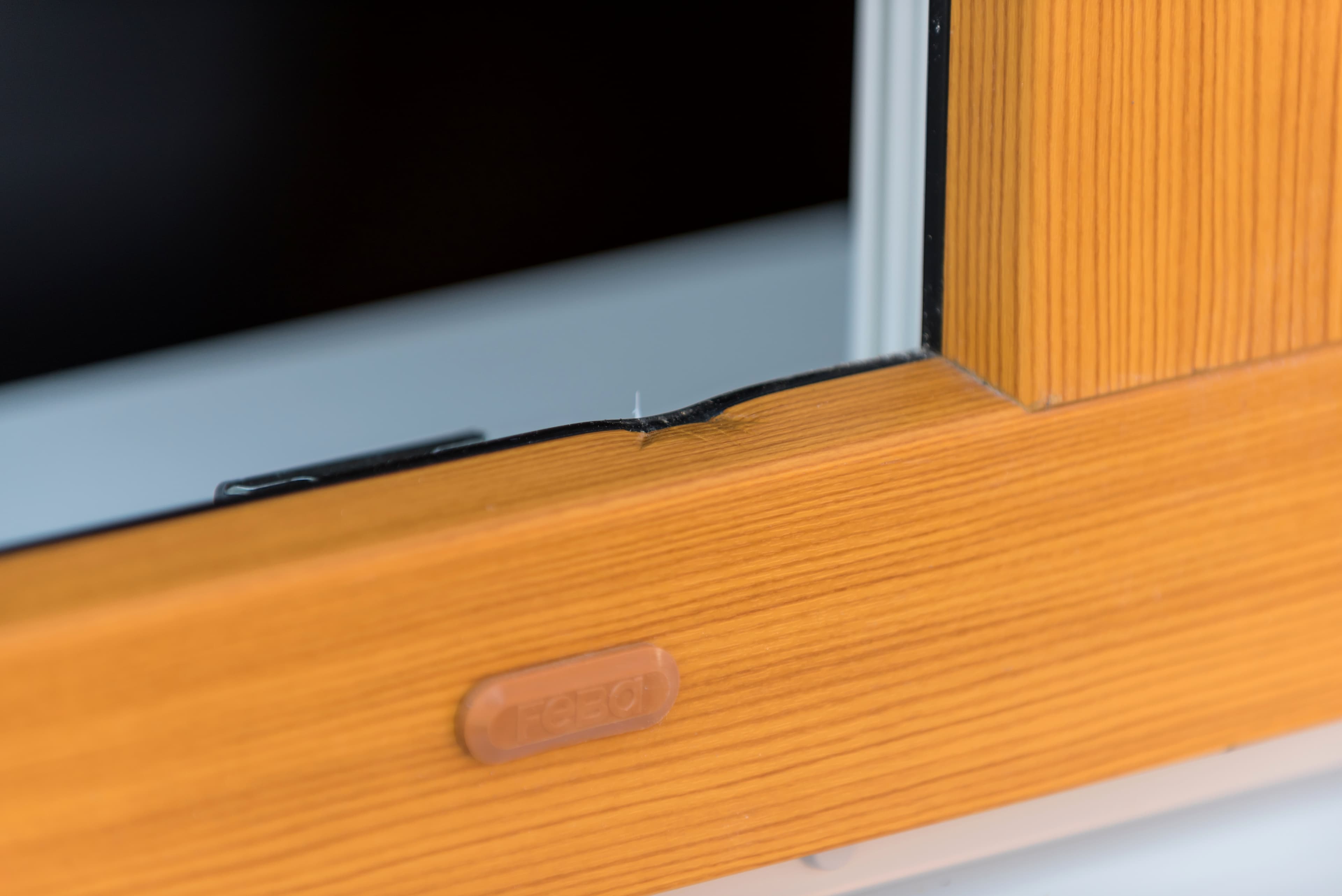 Eine Nahaufnahme eines hölzernen Fensterrahmens mit einem sichtbaren Riss im Glas in der Nähe des Randes. Der Riss ist klein, aber auffällig, und ein braunes Etikett ist am Rahmen befestigt.