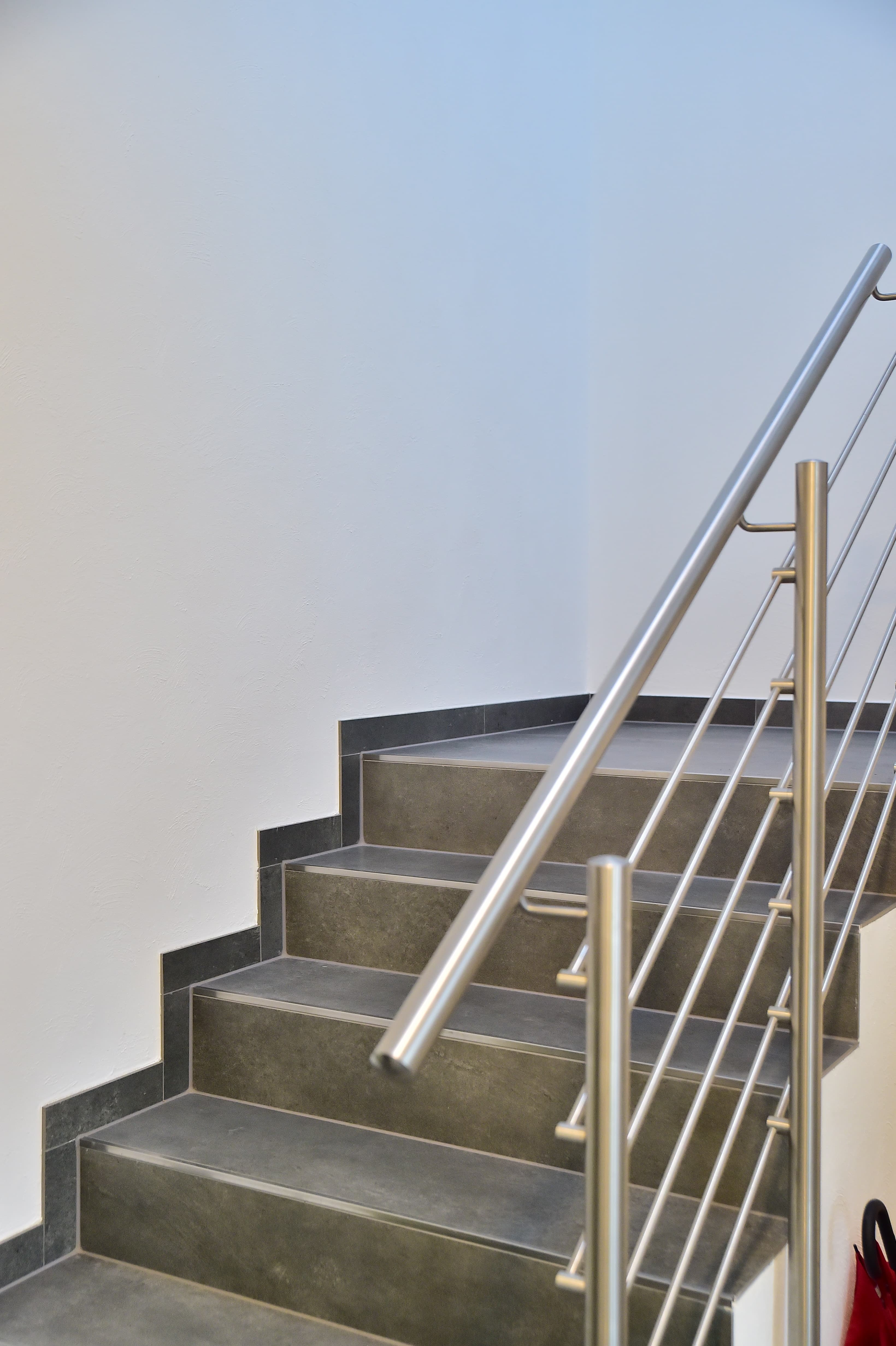 Eine moderne Innentreppe mit dunkelgrauen Stufen und einem Handlauf aus Edelstahl vor einer weißen Wand. Das Design ist minimalistisch und sauber, mit geometrischen Linien und ohne sichtbare Verzierungen.