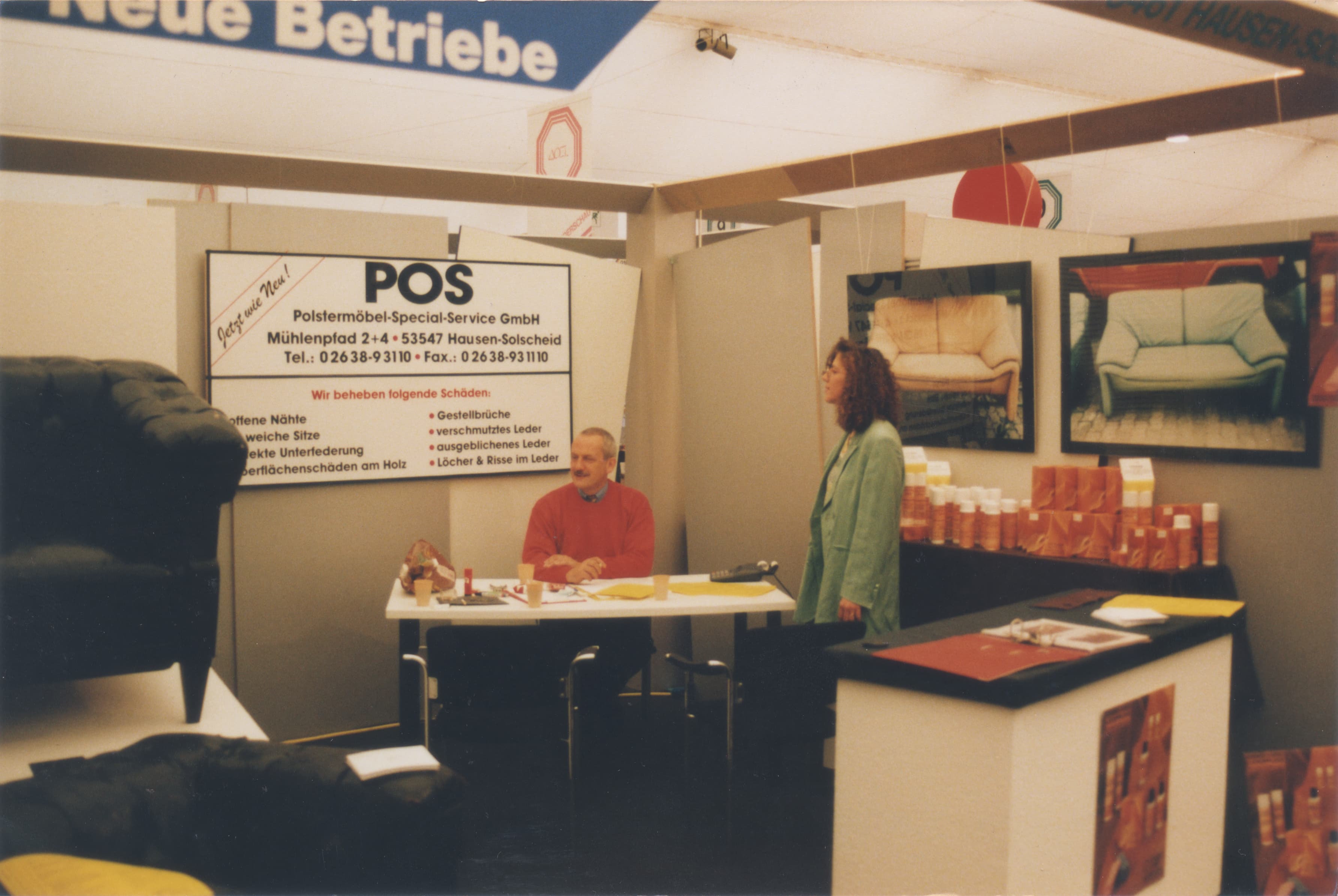 Ein Mann, der an einem Stand sitzt, und eine Frau, die neben ihm steht, auf einer Messe. Hinter ihnen ist ein Schild der POS Polstermöbel-Spezial Service GmbH mit Kontaktdaten und Abbildungen von Polstermöbeln zu sehen.