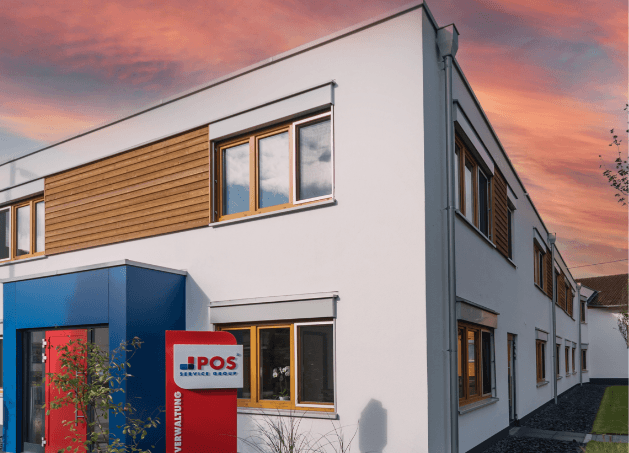 Modernes weißes Bürogebäude mit hölzernen Fensterakzenten und einem rot-blau-weißen POS Service GmbH-Schild am Eingang vor einem dramatischen Sonnenuntergangshimmel.