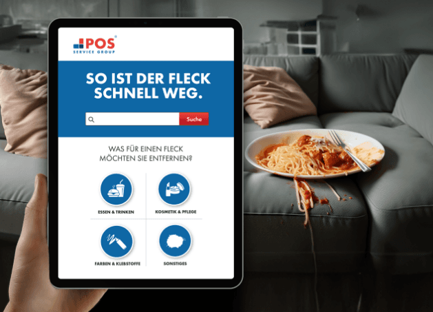 A hand holds a tablet displaying a stain removal website in German, while in the background, a plate of spilled spaghetti and sauce stains a gray couch.