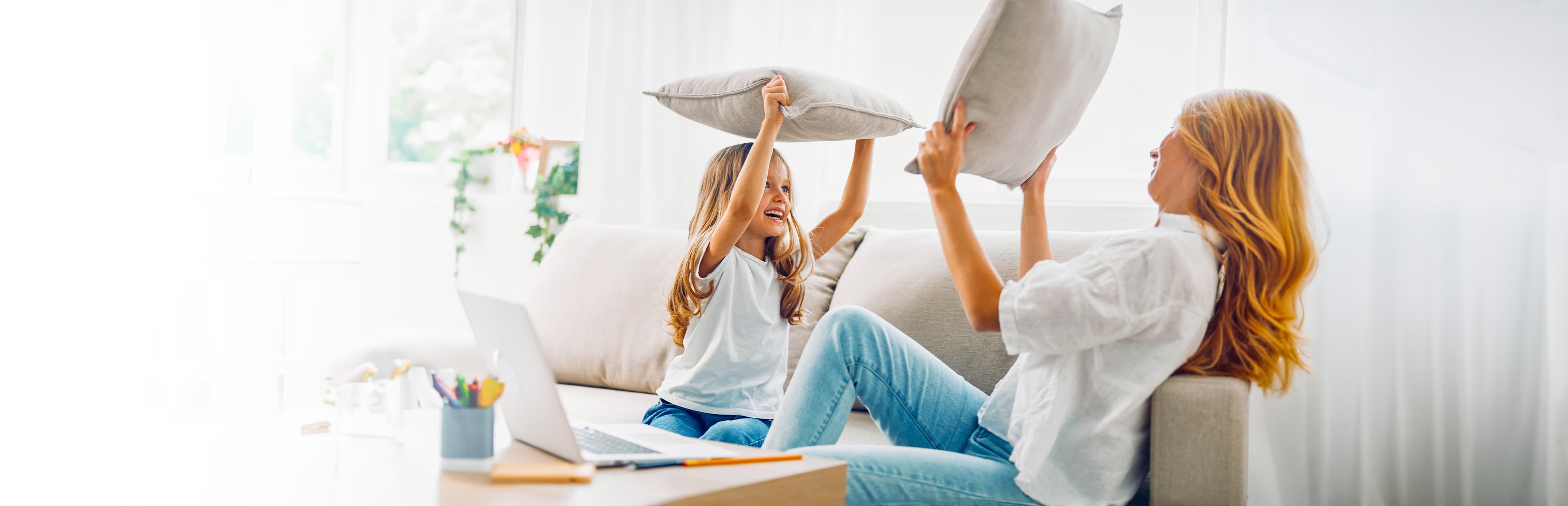 Eine Frau und ein junges Mädchen liefern sich eine Kissenschlacht auf einer hellen, gemütlichen Couch. Beide lächeln und tragen legere Kleidung. Ein Laptop und Buntstifte liegen auf dem Tisch vor ihnen.