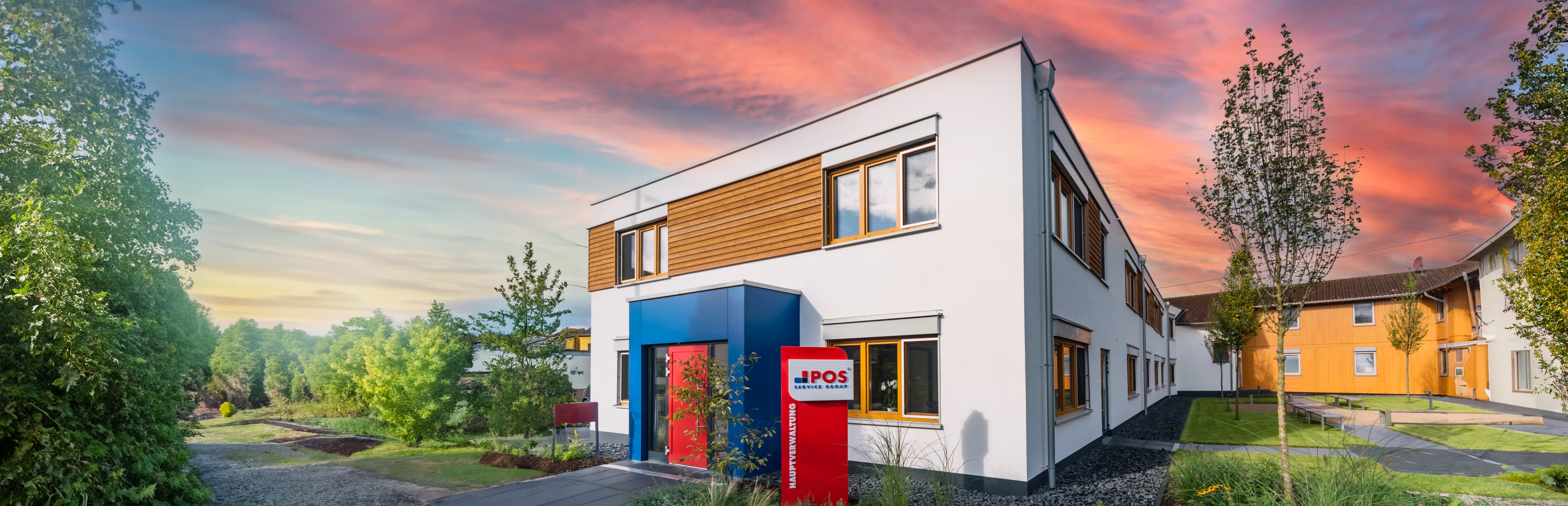 Een modern gebouw van twee verdiepingen met witte en houten lambrisering ligt midden in het groen bij zonsondergang, met een rood bord met de tekst IPOS bij de ingang en een kleurrijke lucht op de achtergrond.