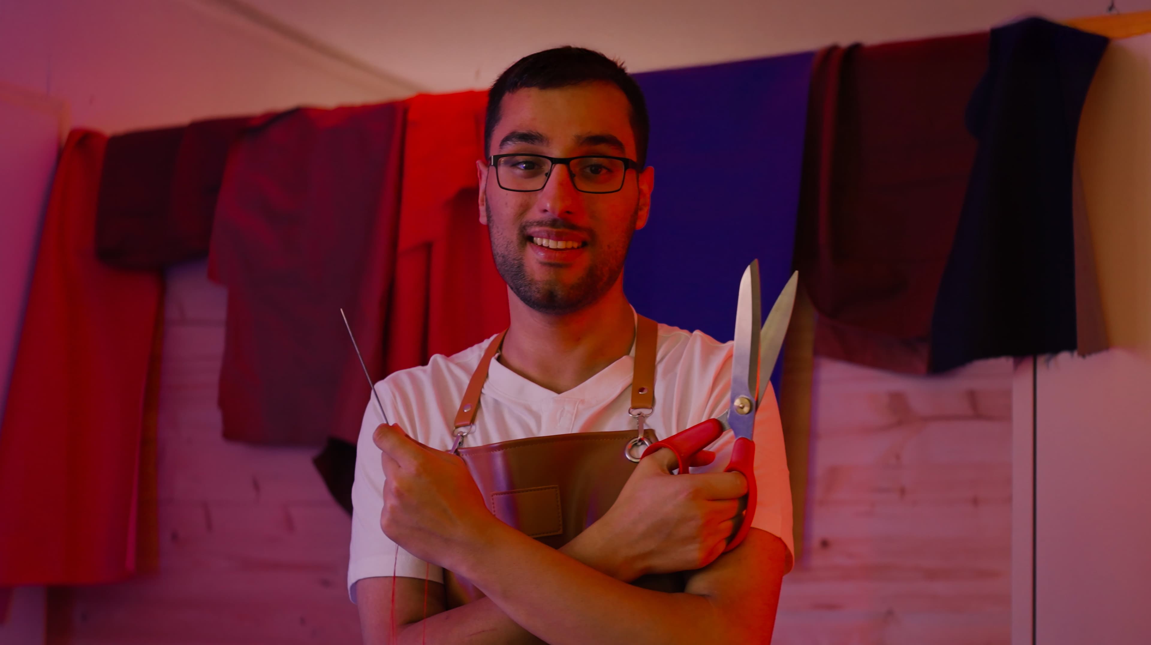 Een man met een bril en een schort staat voor kleurrijke gedrapeerde stoffen, glimlacht en houdt een grote schaar en een stuk gereedschap in zijn gekruiste armen.