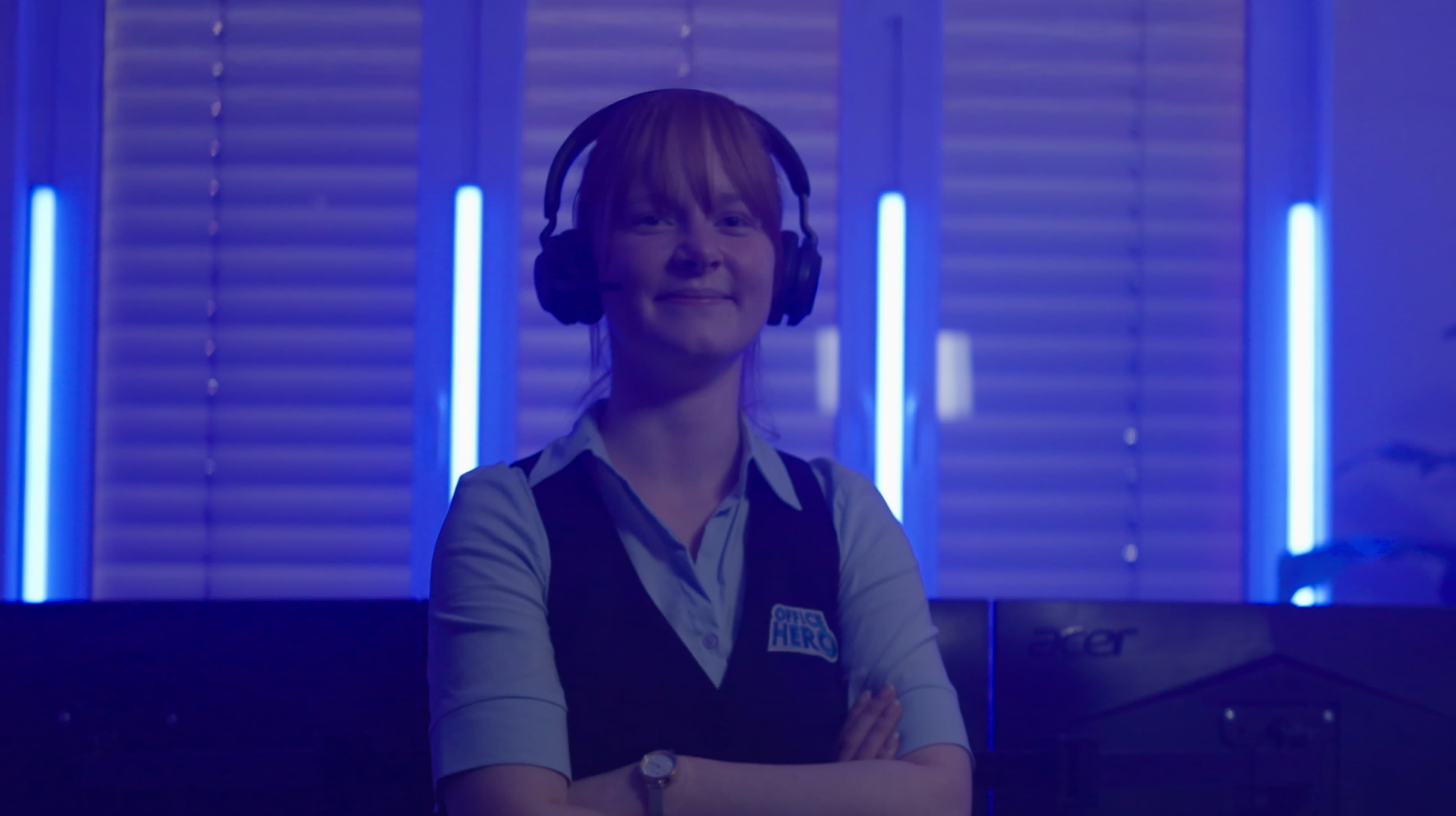 Een glimlachende vrouw met een headset en een blauw vest met een HIRE HERO badge staat met haar armen over elkaar in een schemerige kamer met blauwe lampen en computerschermen.
