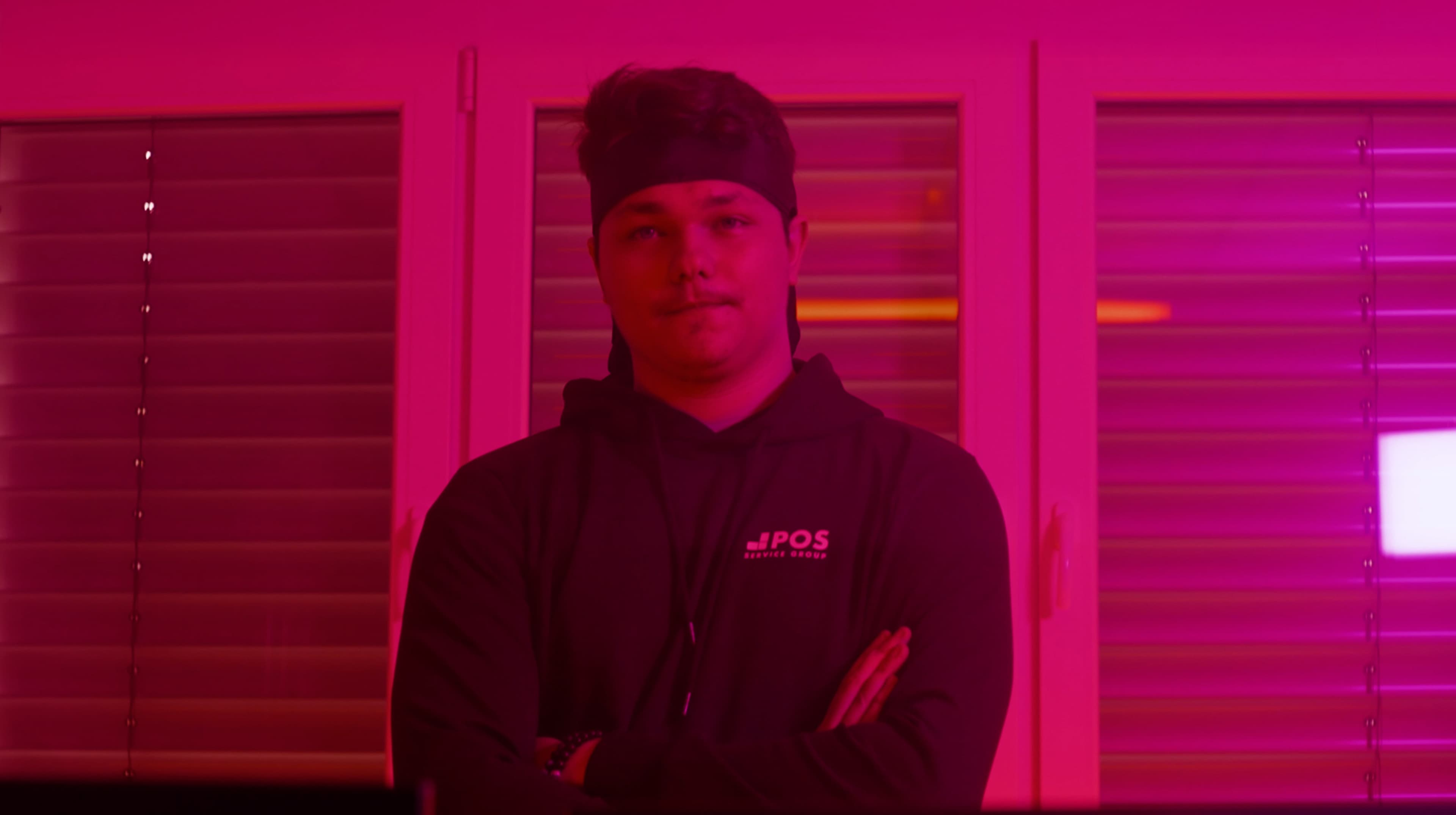 Een persoon met een hoofdband en een donkere capuchon staat met gekruiste armen voor gesloten jaloezieën, verlicht door roze-paars licht.