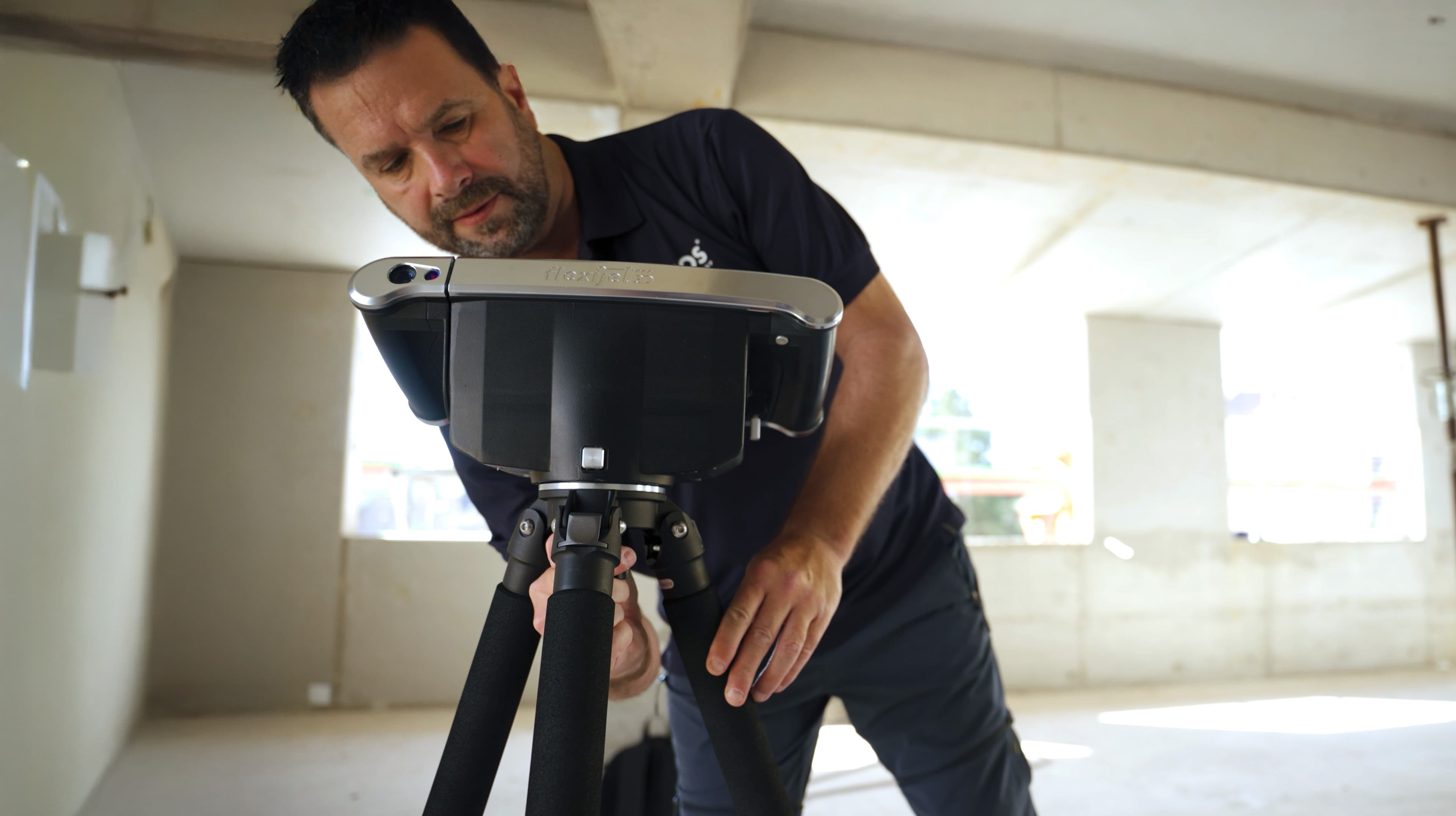 Ein Mann justiert ein auf einem Stativ montiertes 3D-Scangerät im Inneren eines unfertigen Gebäudes mit Betonwänden und großen Fenstern.