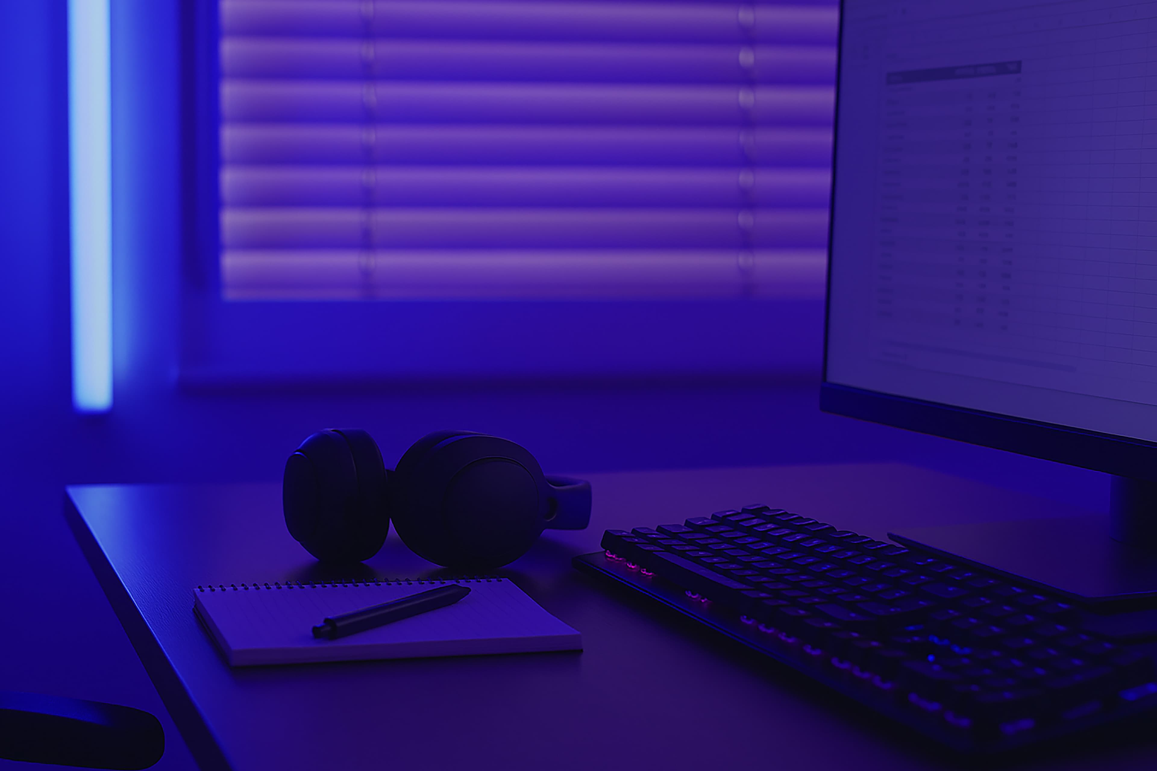 A desk with a keyboard, computer monitor, notepad, pen, and headphones, lit by purple-blue ambient lighting from a window with closed blinds in the background.