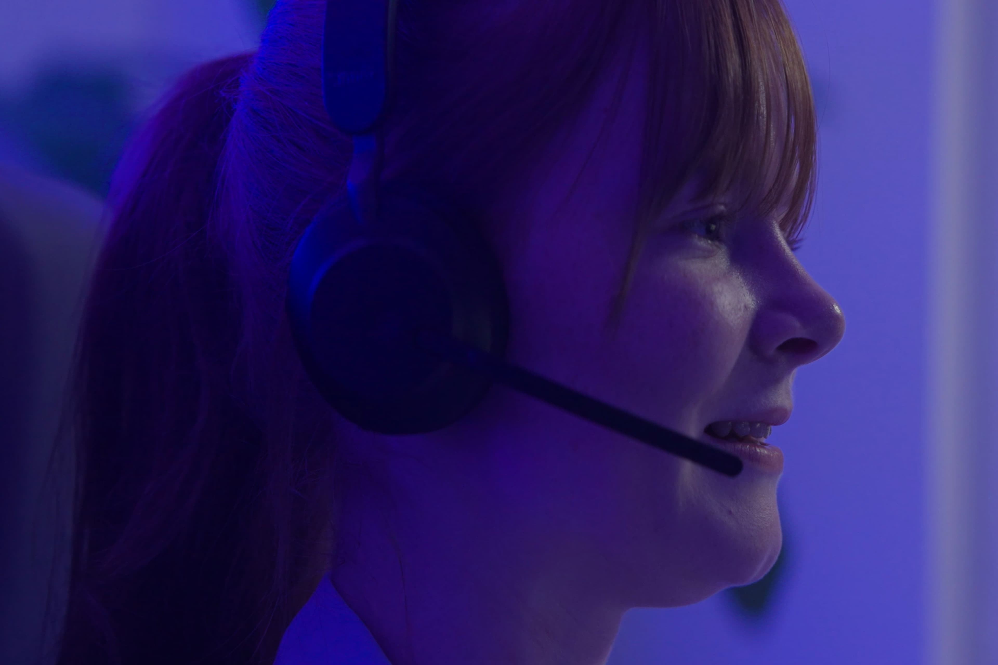 A close-up of a smiling person with red hair wearing a headset microphone, bathed in blue lighting, suggesting they are engaged in a conversation or gaming.