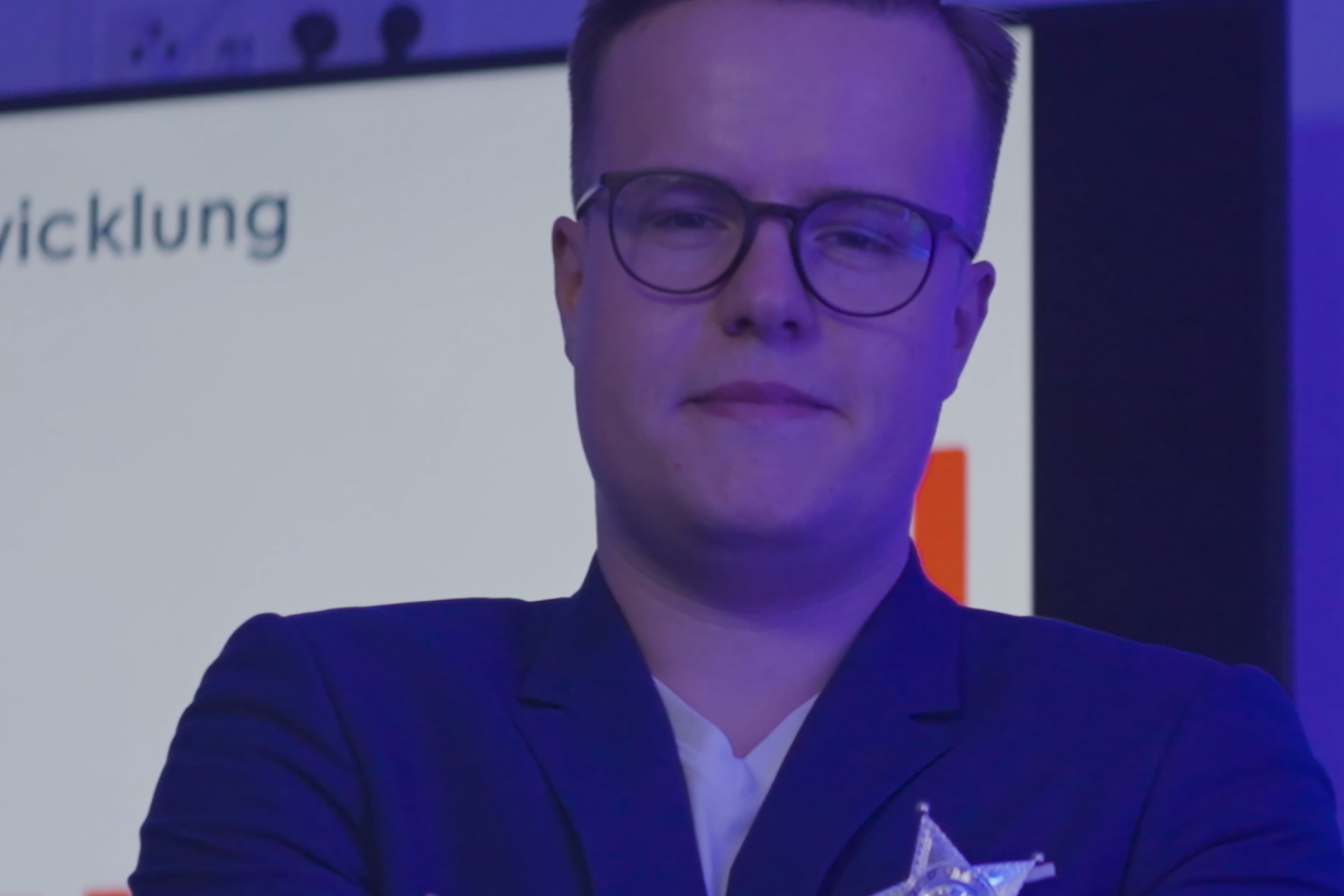 Une personne aux cheveux courts et aux lunettes se tient devant un écran de présentation flou, vêtue d'un blazer sombre et d'une chemise blanche, sous un éclairage bleu violet.