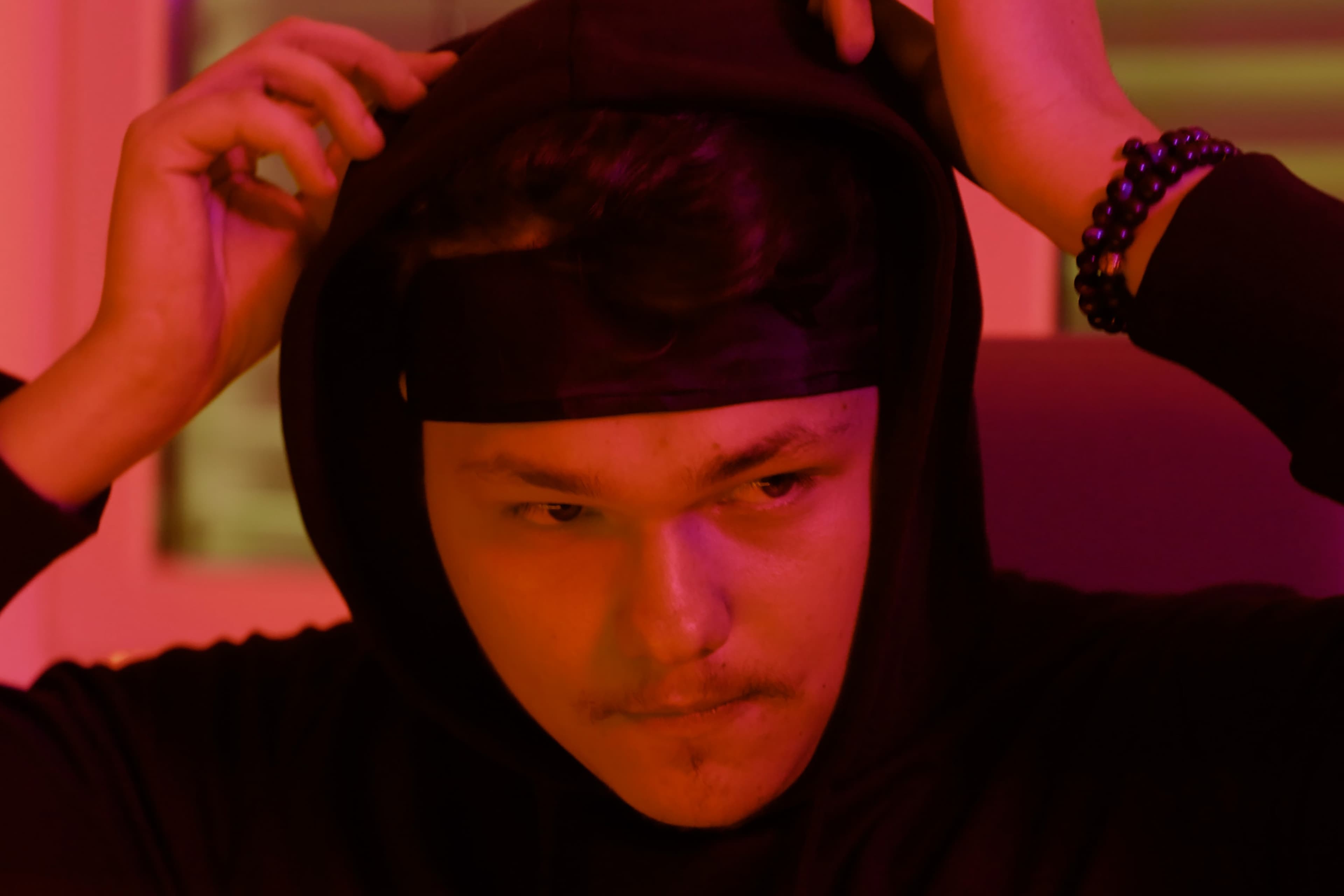A young person with a faint mustache pulls a black hood over their head, illuminated by moody red and purple lighting. They wear a black wristband and look focused, gazing slightly downward.