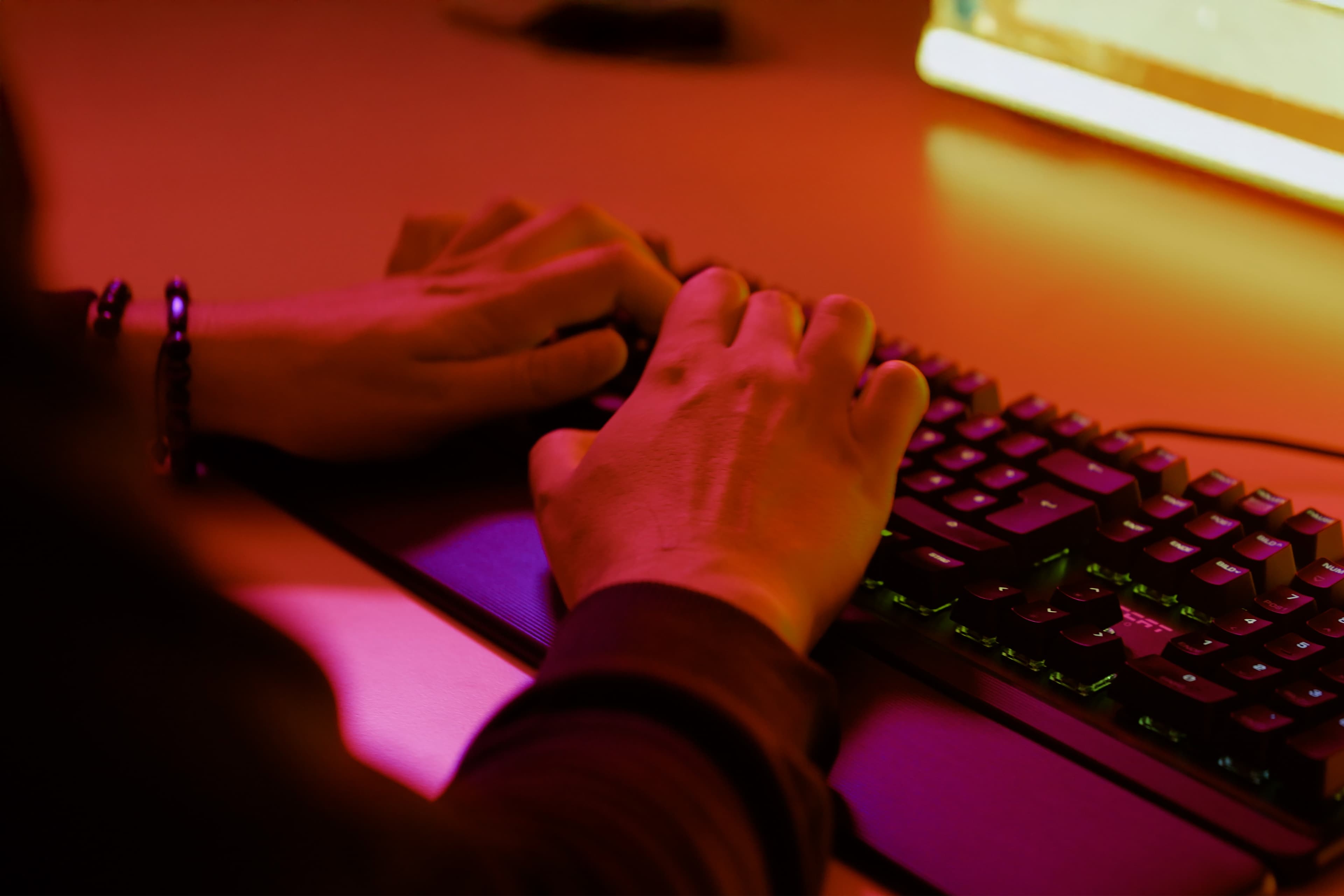 Close-up of hands typing on a keyboard, illuminated by warm red and yellow lighting from a computer screen on a desk.
