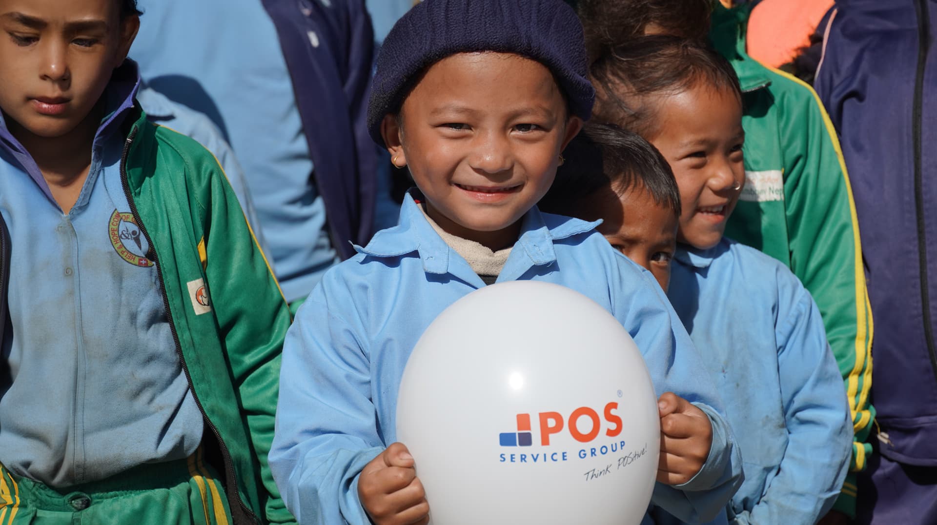 Ein lächelndes kleines Kind in blauer Uniform und Strickmütze hält einen weißen Luftballon mit dem Logo der POS Service Group. Andere Kinder in bunten Uniformen stehen neben ihm, alle draußen im Sonnenlicht.