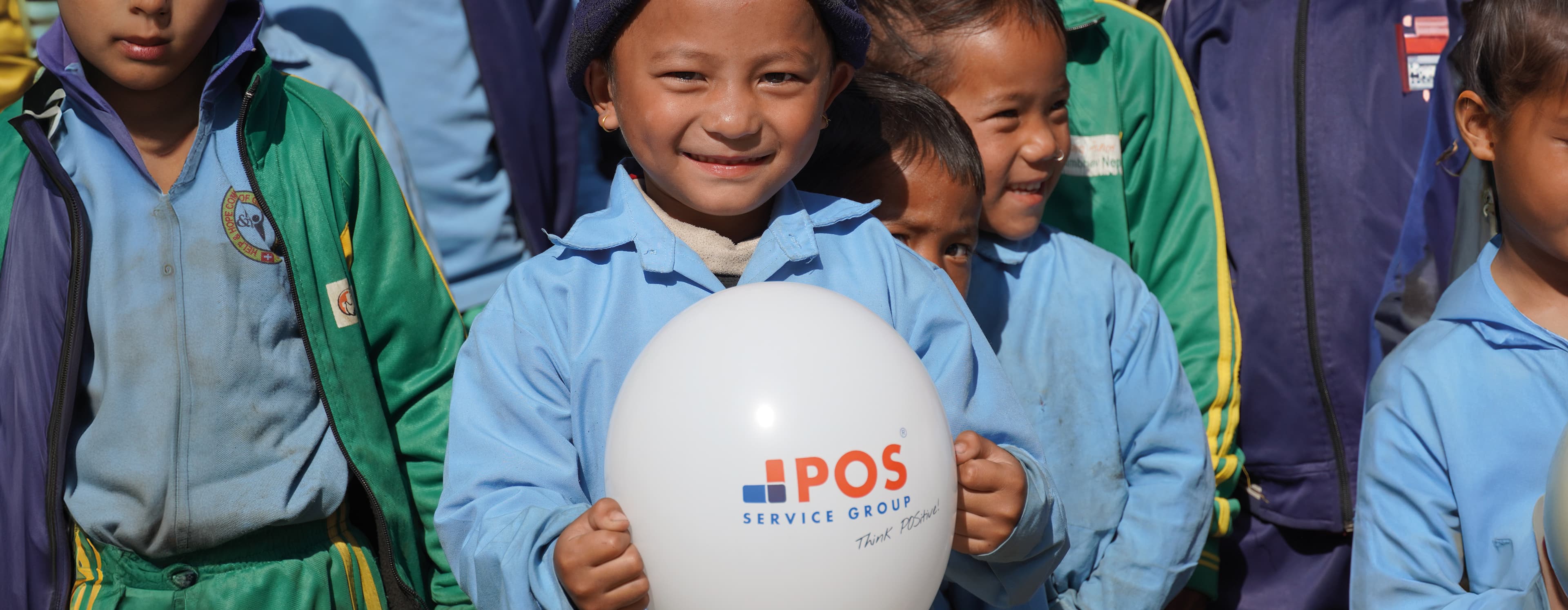 A group of young children in blue and green school uniforms stand together outdoors. One child in front smiles while holding a white balloon with the POS Service Group logo.