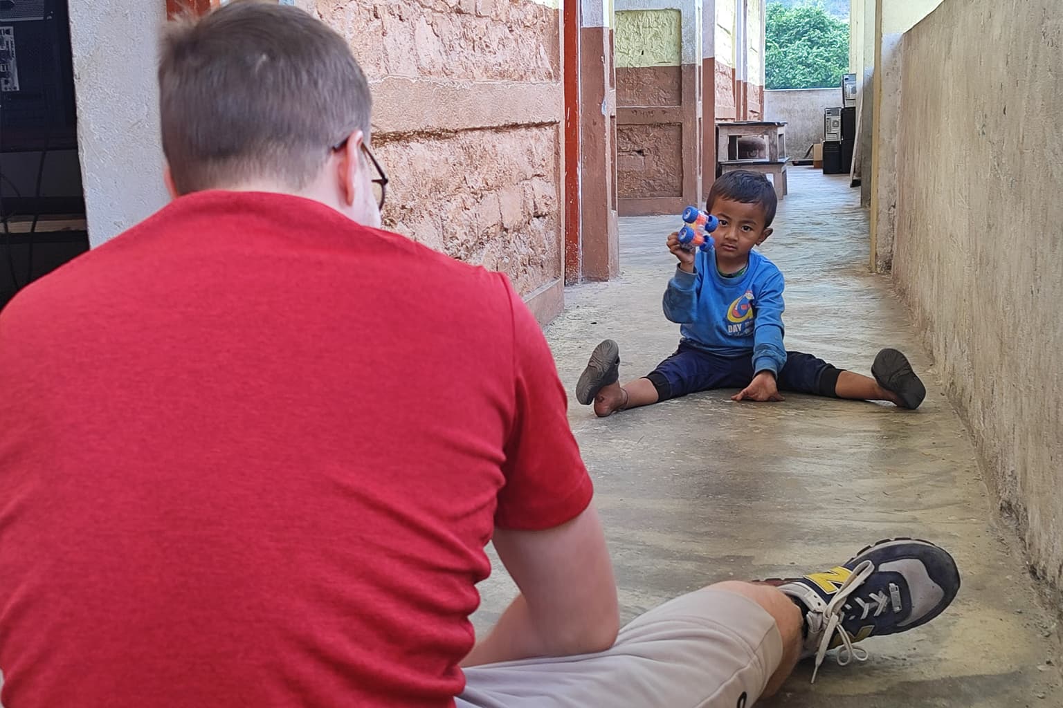 A young boy sits on the ground with legs spread, holding up a blue toy and smiling at a man in a red shirt who sits facing him in a corridor with stone walls.