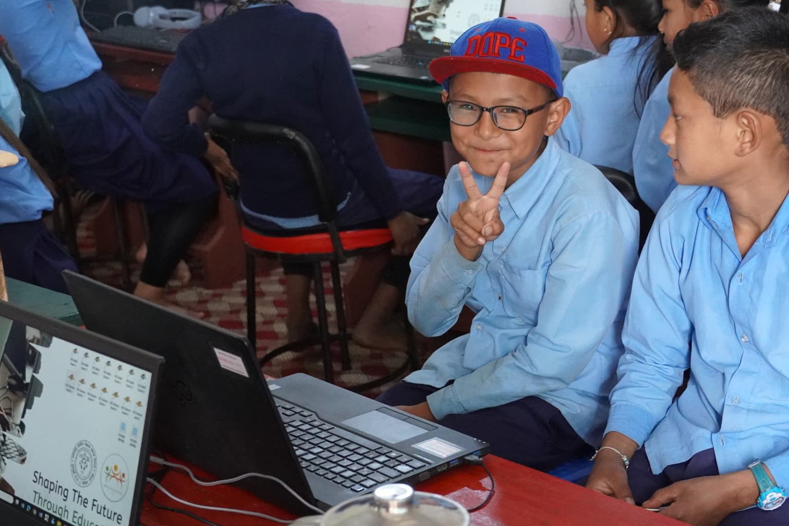 A boy in a blue shirt and red cap sits next to another student, smiling and making a peace sign in front of a laptop in a classroom setting, with other students in the background.