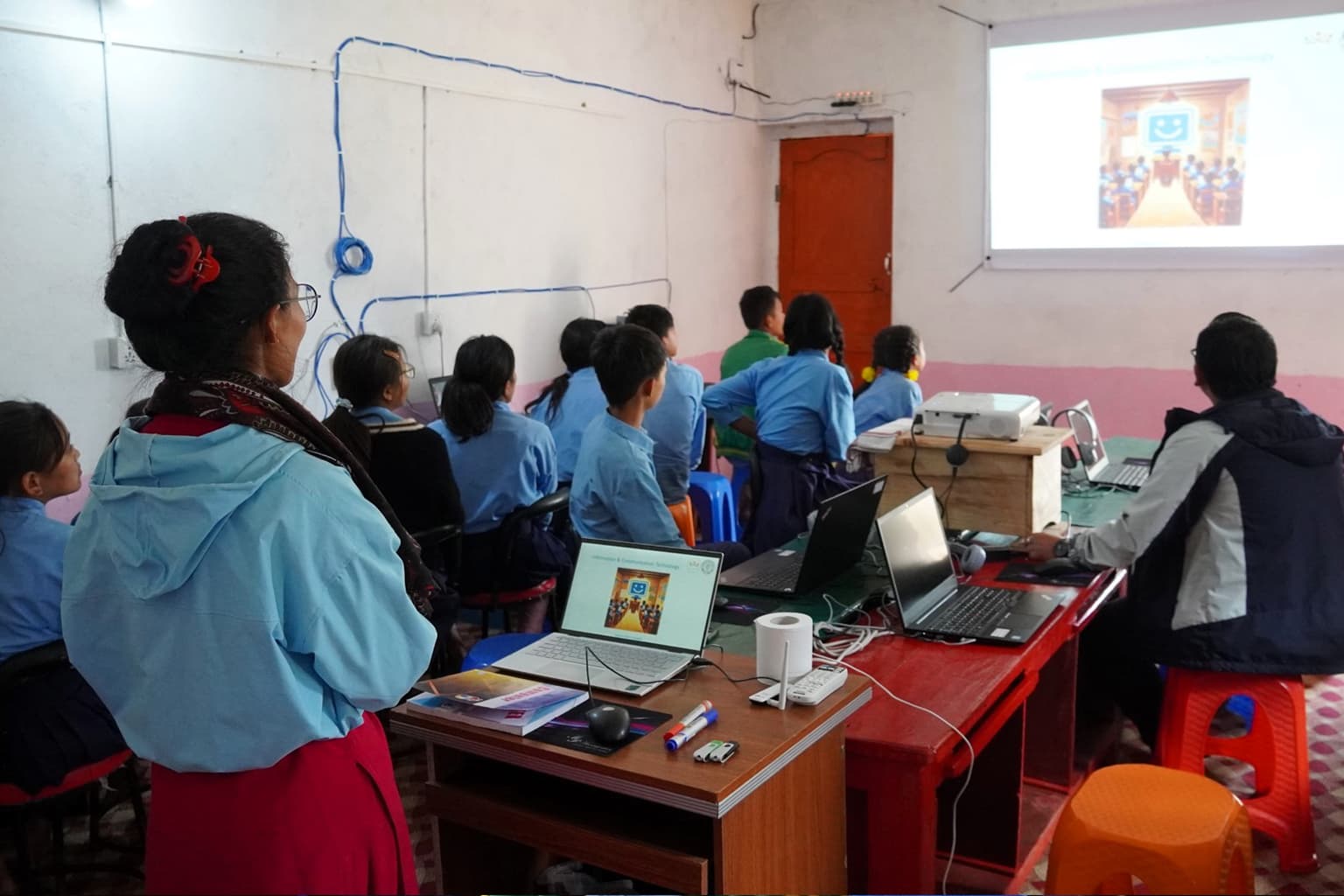 A classroom with students in blue uniforms seated at desks, watching a presentation on a screen. Two adults supervise while laptops and educational materials are on the tables. The room has white walls and visible wiring.