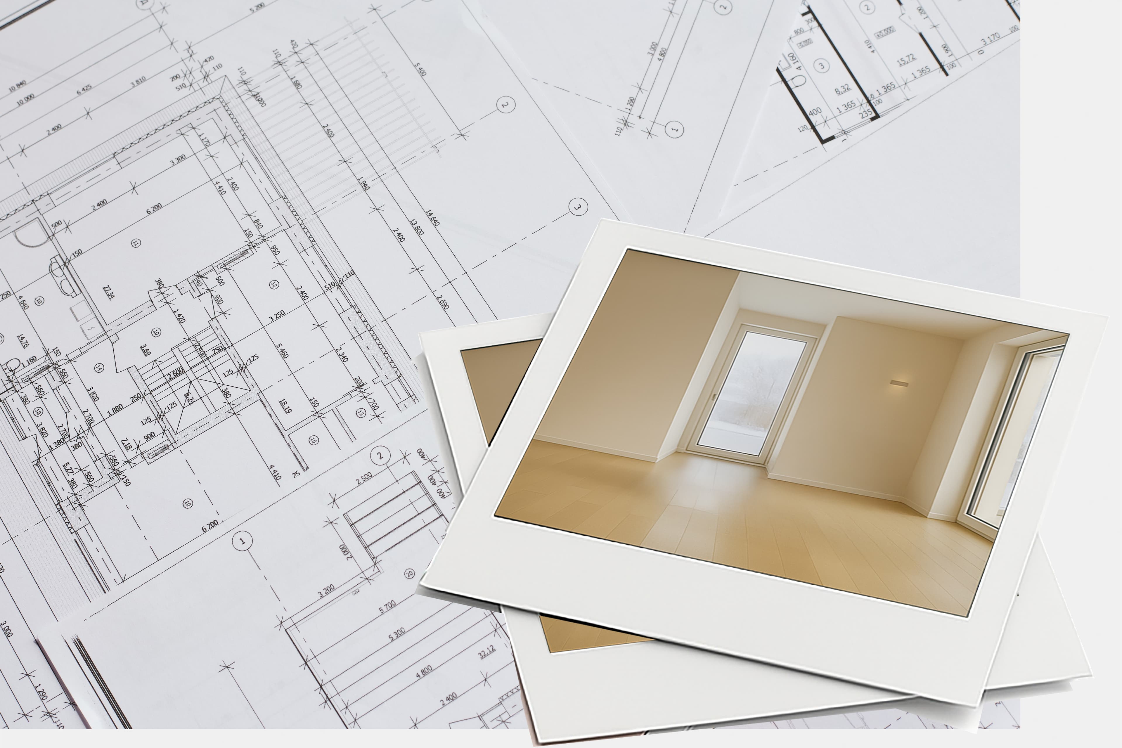 Two printed photos of an empty, bright room with wooden floors and large window are placed on top of architectural blueprints featuring detailed measurements and drawings.