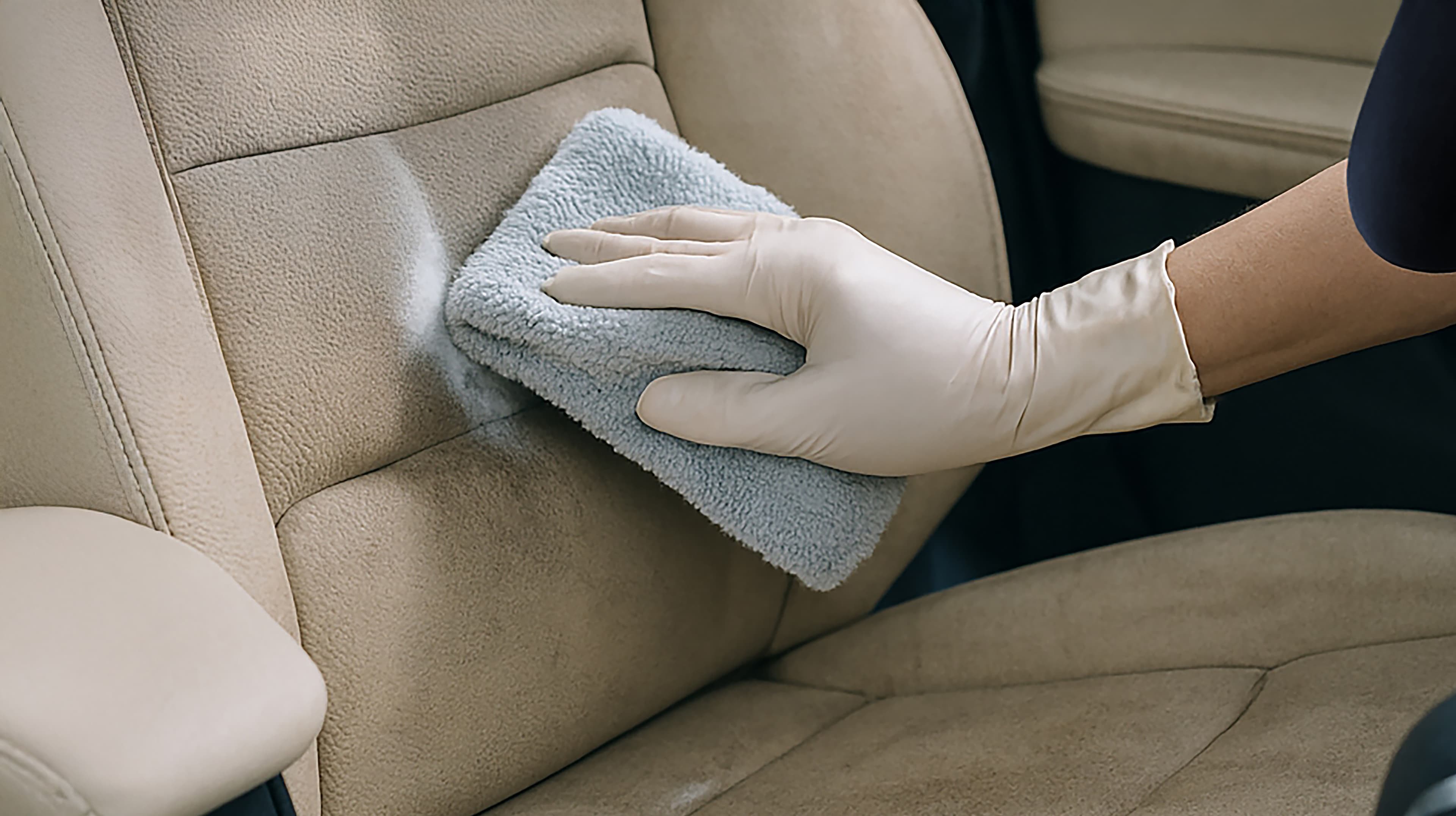 Een persoon die een latexhandschoen draagt, reinigt een beige autostoel met een lichtblauwe microvezeldoek en veegt vuil of vlekken weg.
