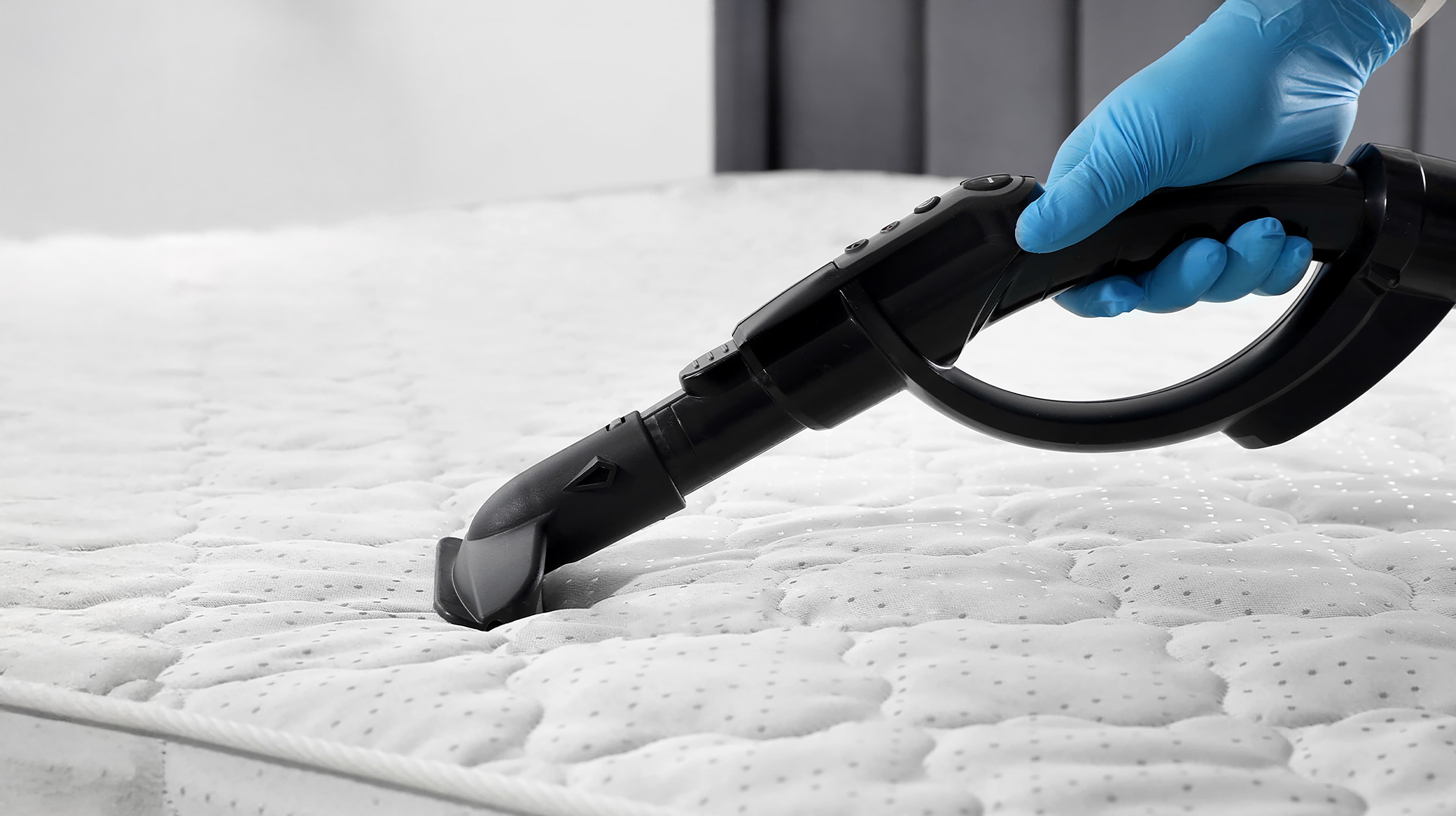 Eine behandschuhte Hand reinigt mit einem Staubsauger eine weiße Matratze, wobei sie sich auf die Entfernung von Staub und Allergenen konzentriert.