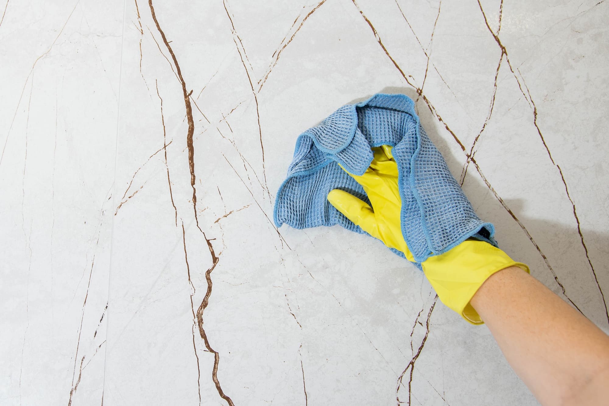 Eine Person mit einem gelben Gummihandschuh reinigt mit einem blauen Lappen eine weiße Fliesenfläche mit braunen Rissen.