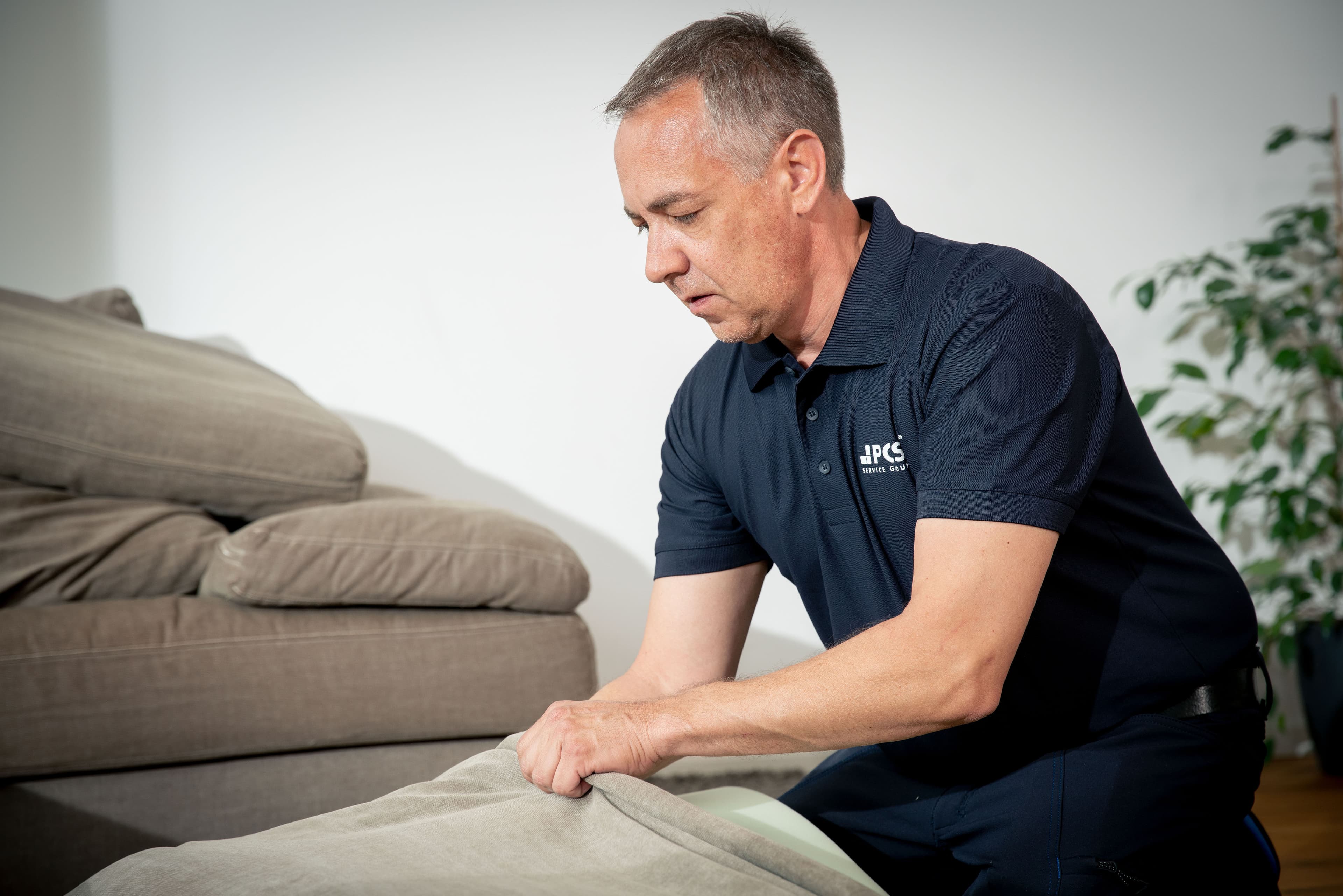 Een man in een marineblauw poloshirt verwijdert een beige kussenhoes van een bank, met een andere bank en een plant zichtbaar op de achtergrond.