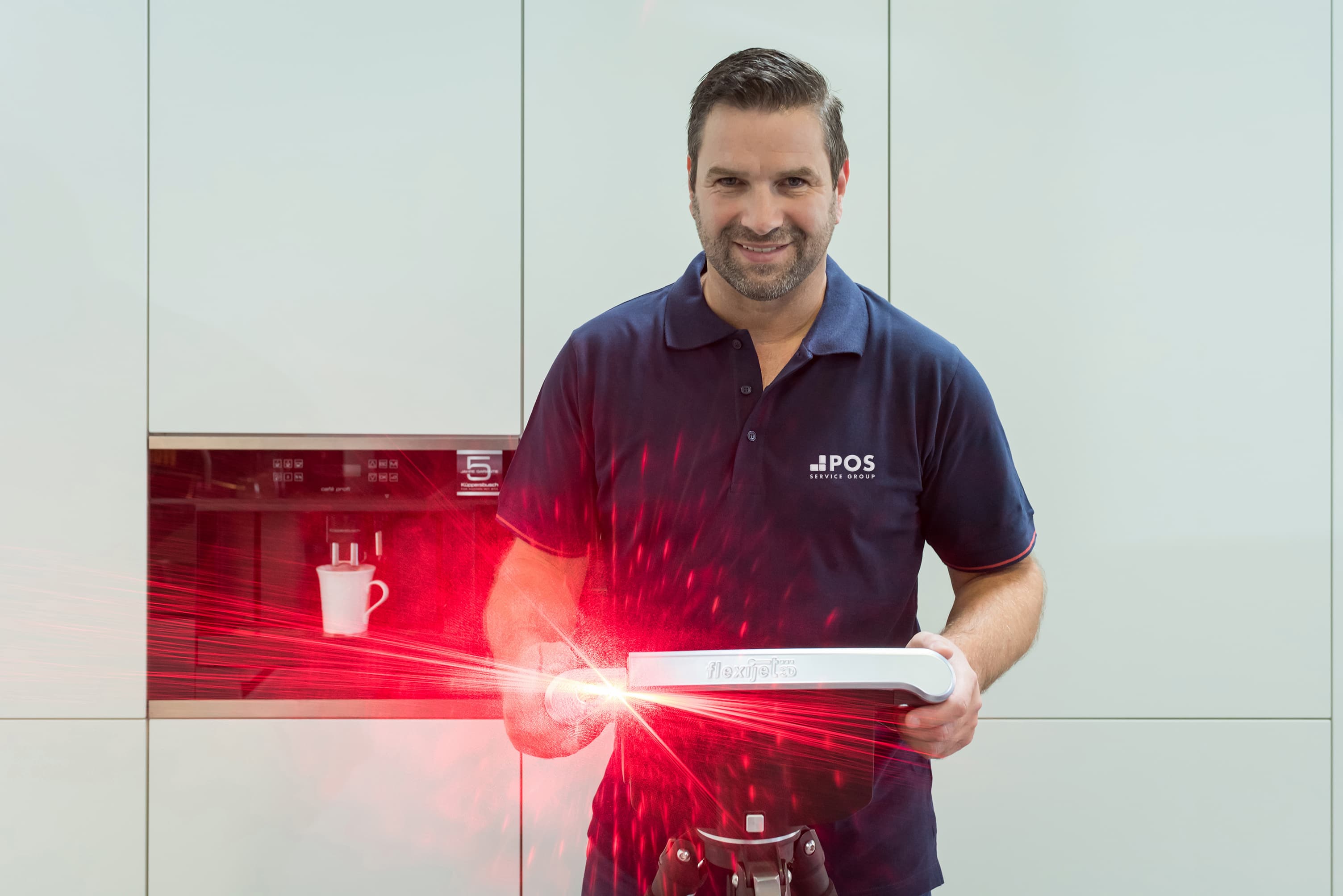 Een man in een marineblauw POS poloshirt glimlacht terwijl hij een apparaat vasthoudt dat een felrood laserlicht uitzendt in een moderne keuken, met een koffiekopje en een machine op de achtergrond.