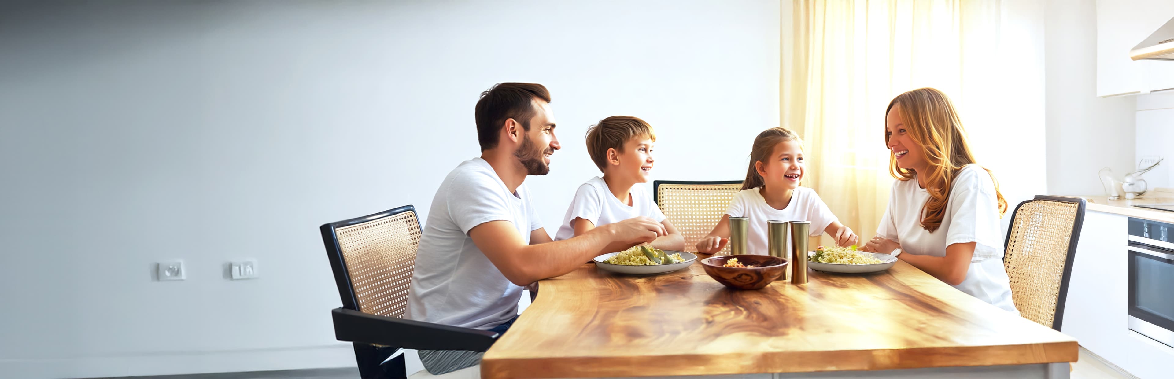 Eine vierköpfige Familie, zwei Erwachsene und zwei Kinder, sitzt an einem hölzernen Esstisch in einer hellen Küche, lächelt und genießt eine gemeinsame Mahlzeit.