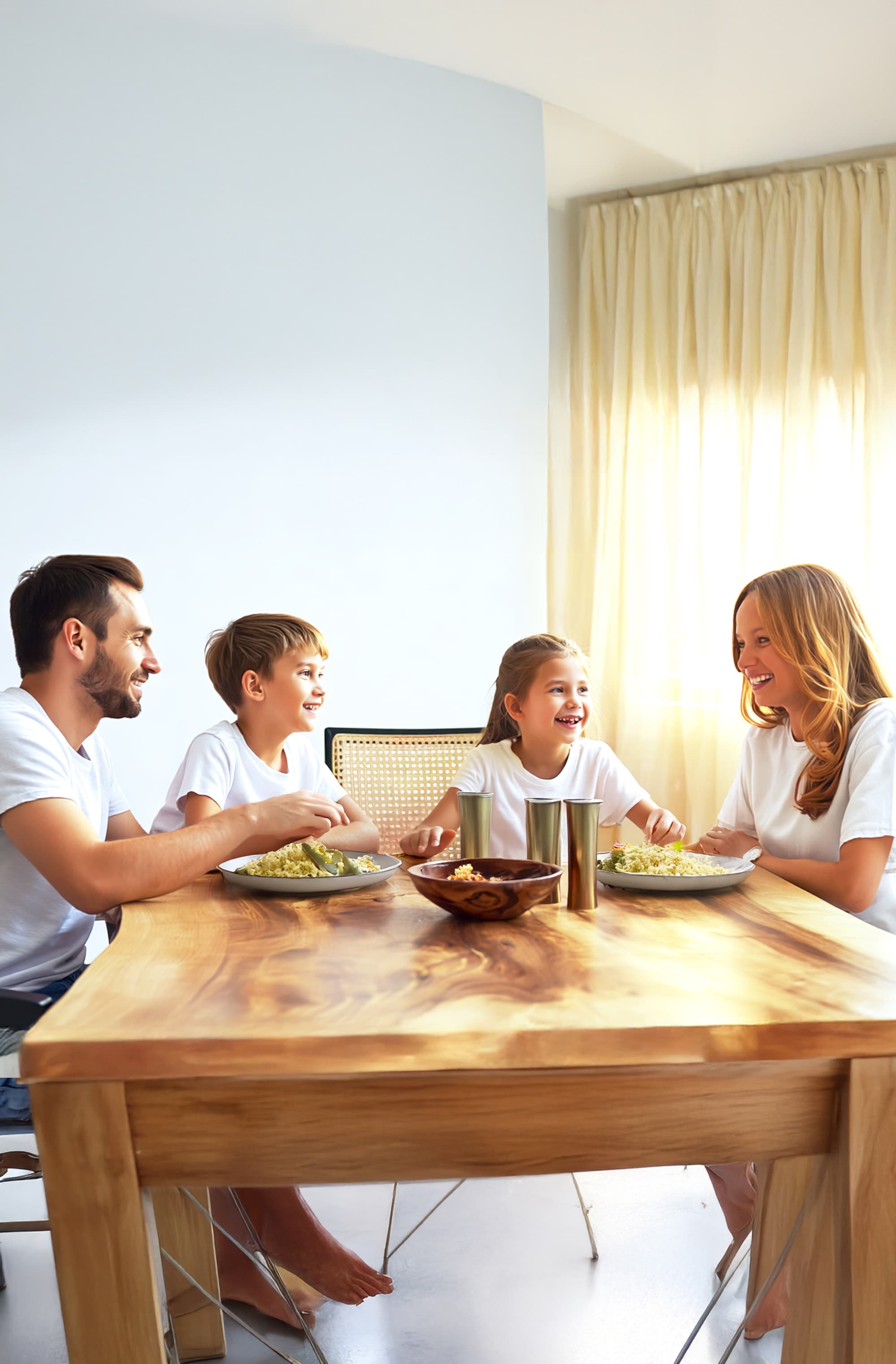 Eine vierköpfige Familie in weißen Hemden sitzt um einen hölzernen Esstisch und isst gemeinsam, lächelt und unterhält sich in einem hellen Raum, in dem das Sonnenlicht durch die Vorhänge fällt.