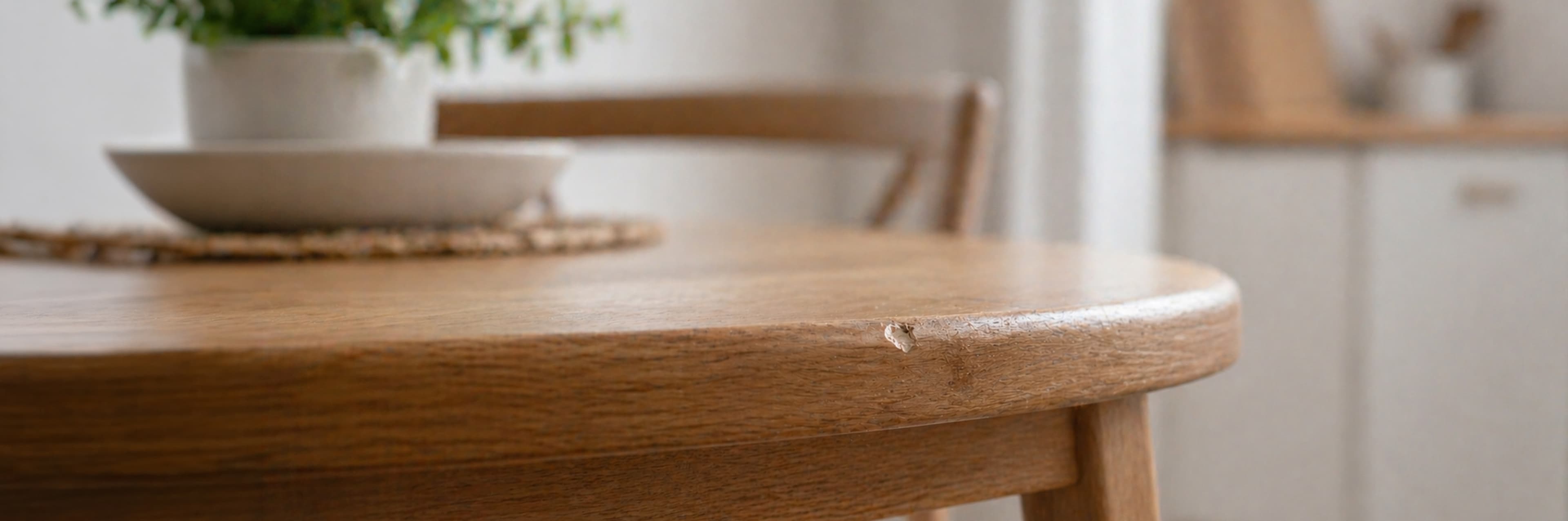 Een close-up van een houten tafel met een zichtbare chip op de rand. Een stoel en een plant in een witte pot op een geweven mat zijn zichtbaar op de achtergrond, samen met een vage keuken.