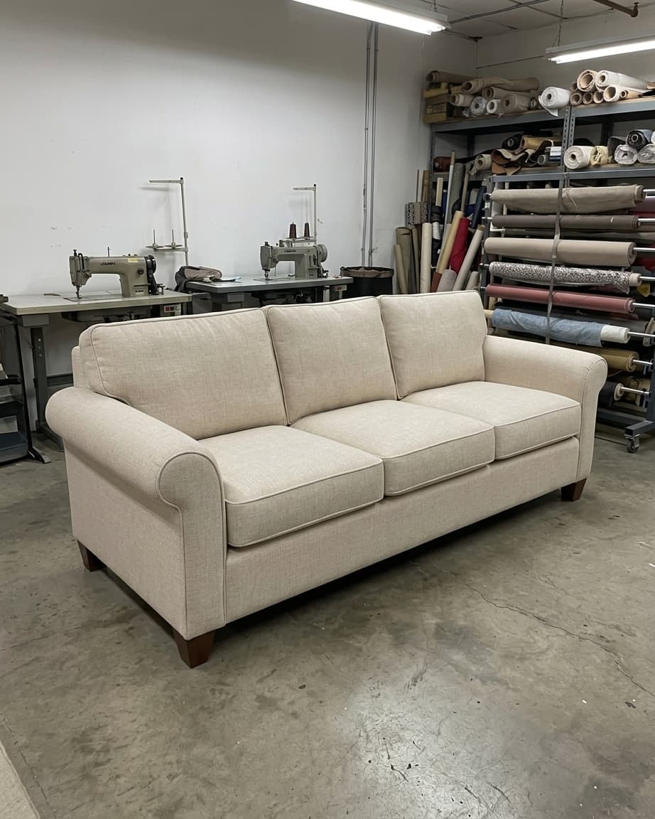 In einer Werkstatt mit Nähmaschinen, Stoffrollen und Regalen im Hintergrund steht unter heller Deckenbeleuchtung ein beiges Dreisitzer-Sofa.